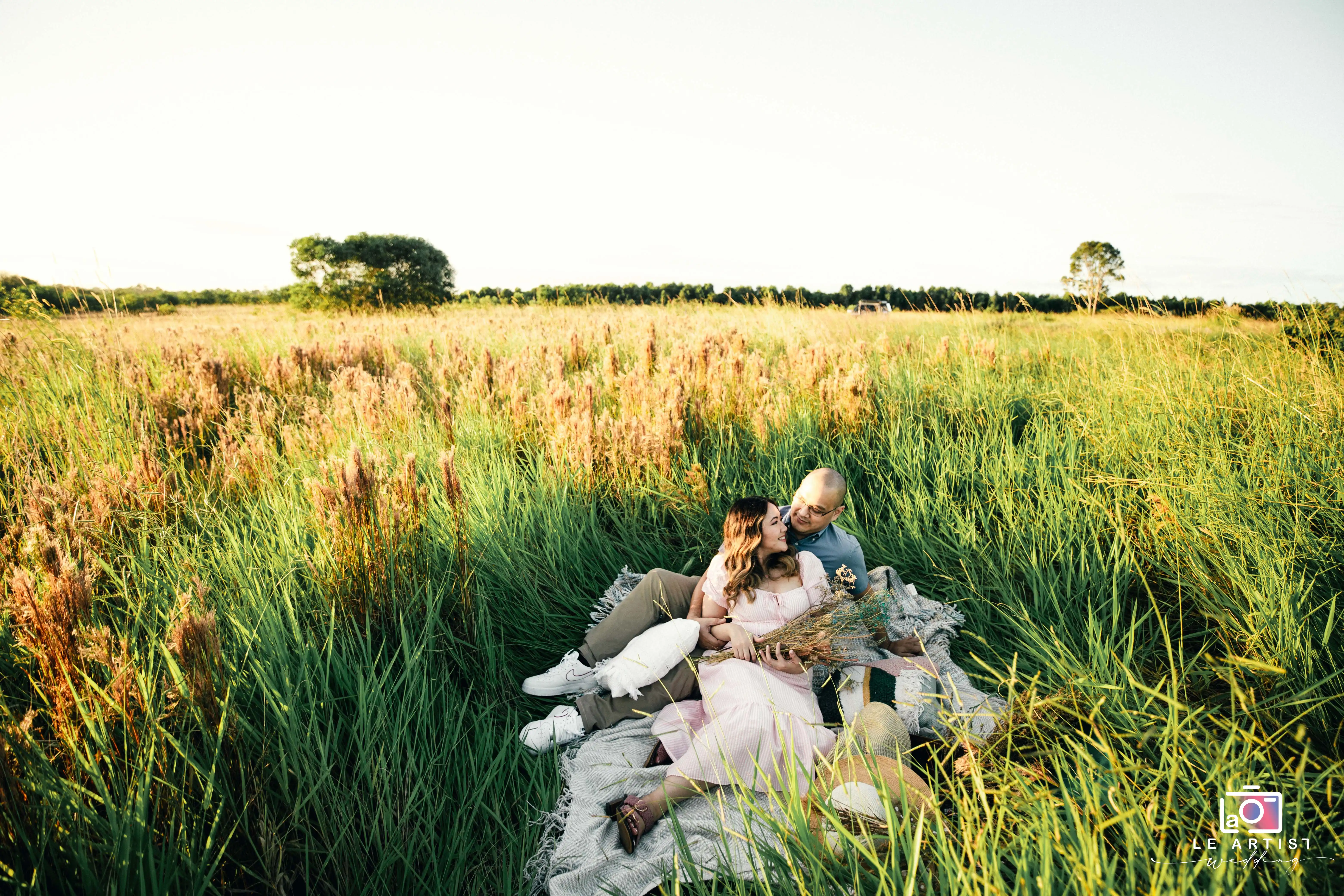 A couple enjoys a serene moment in a sunlit field, surrounded by tall grasses and wildflowers, capturing the essence of romance and nature. Le Artist Wedding