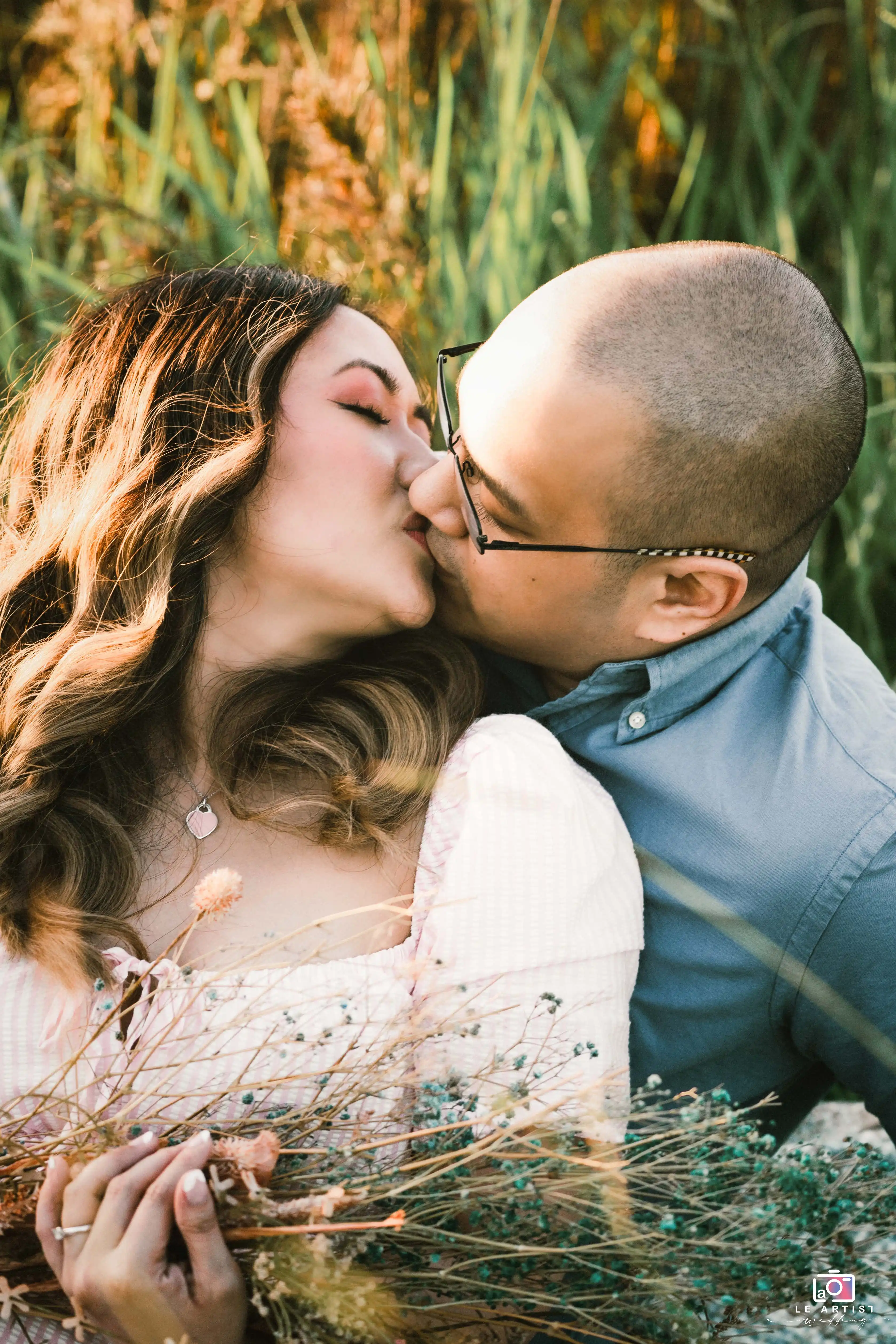 A couple shares a tender kiss in a sunlit field, surrounded by tall grasses and holding a bouquet of wildflowers. Le Artist Wedding