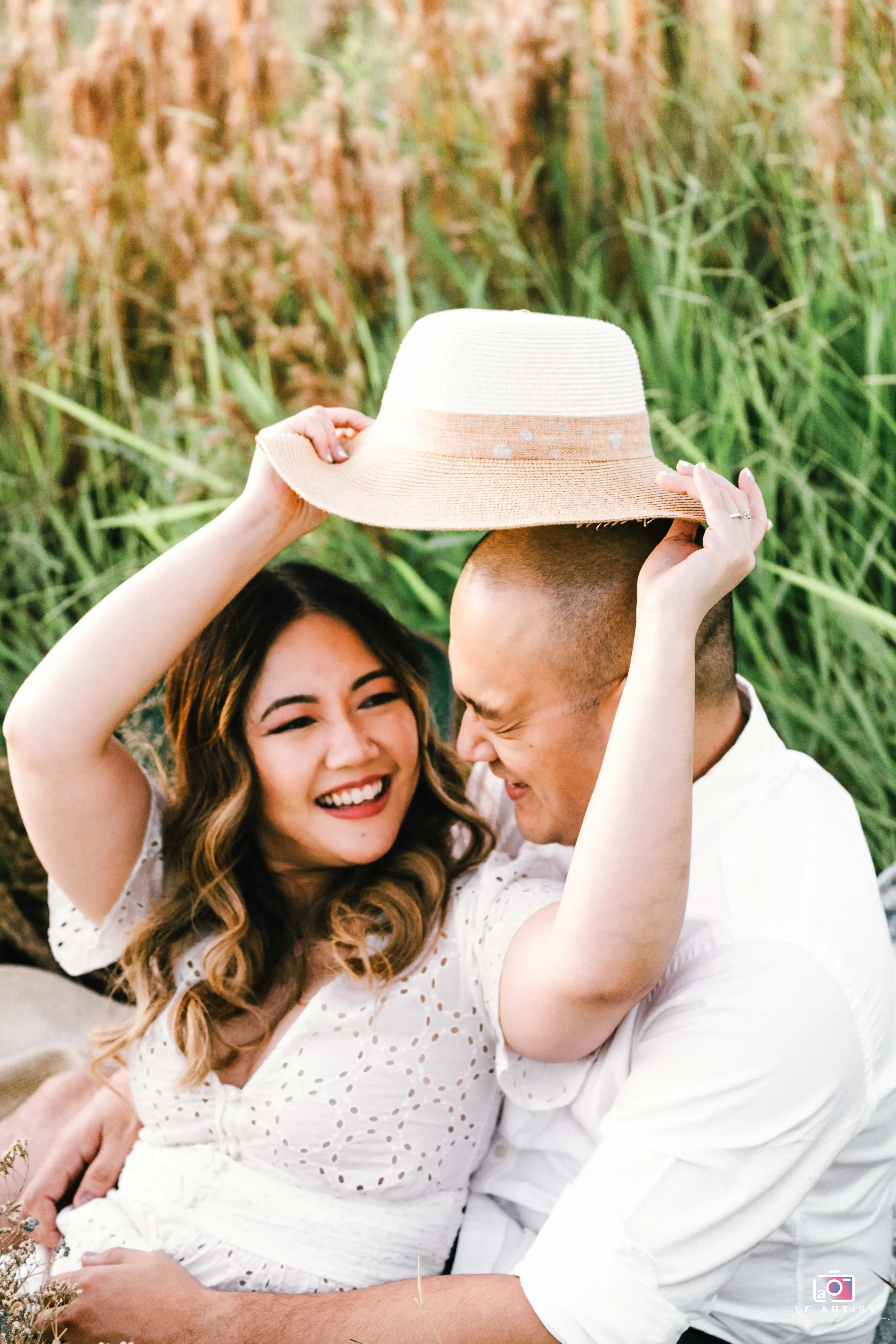 A joyful couple shares a lighthearted moment in nature, with the bride playfully holding her hat over the groom's head. Le Artist Wedding