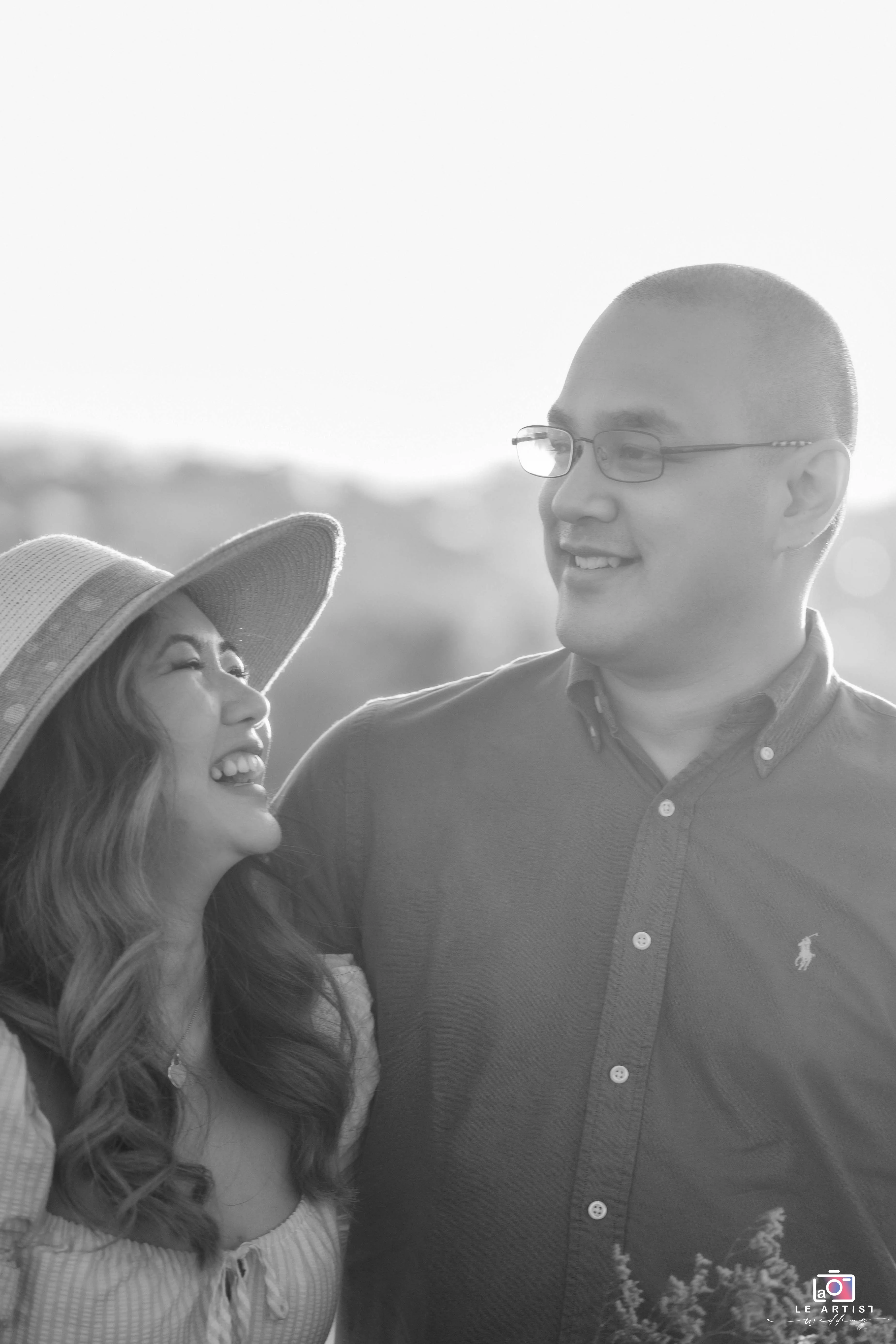A joyful couple shares a moment of laughter during their engagement shoot, capturing their love in a sunlit background. Le Artist Wedding