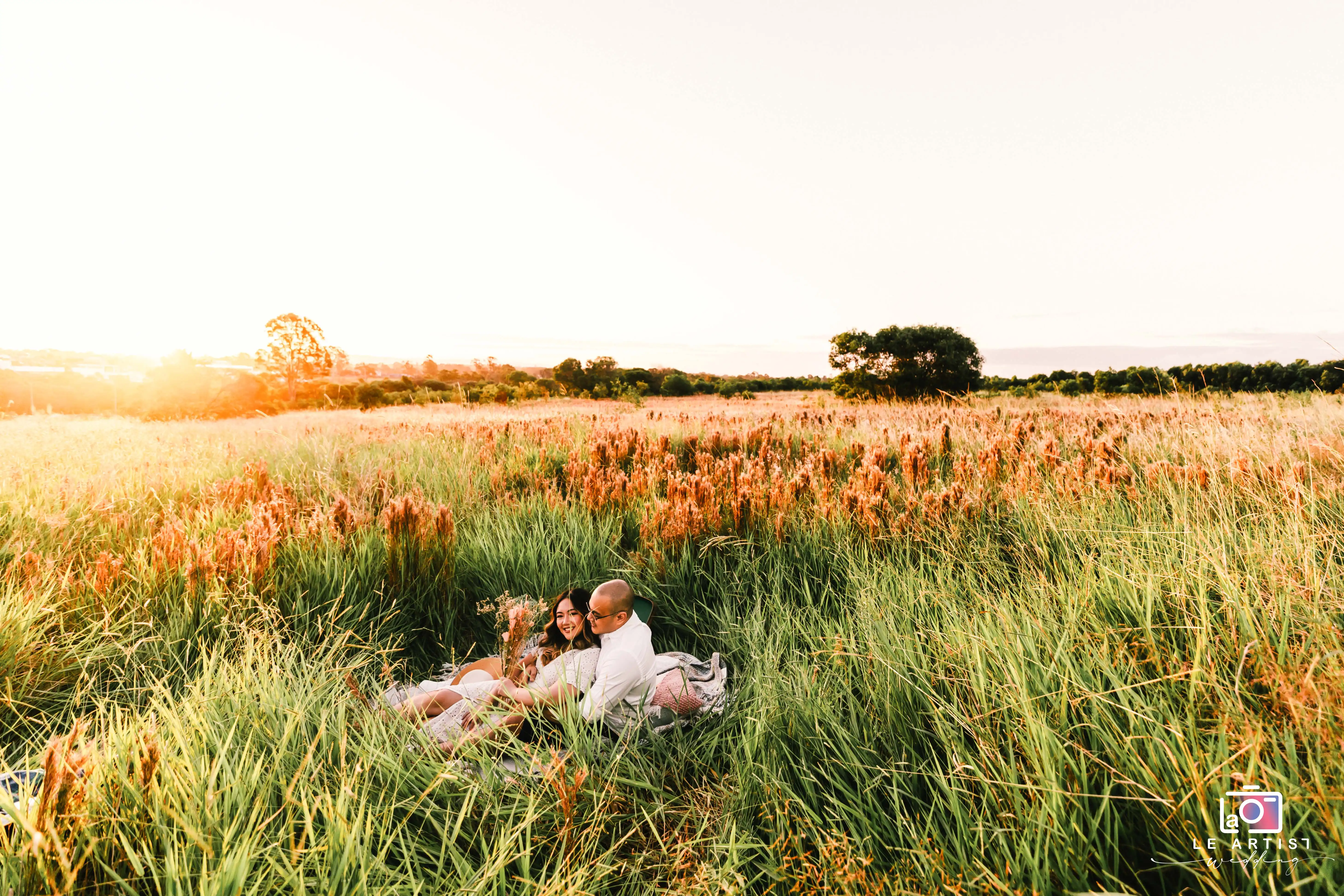 A couple relaxes in a sunlit field, enjoying a peaceful moment during their wedding day. Le Artist Wedding