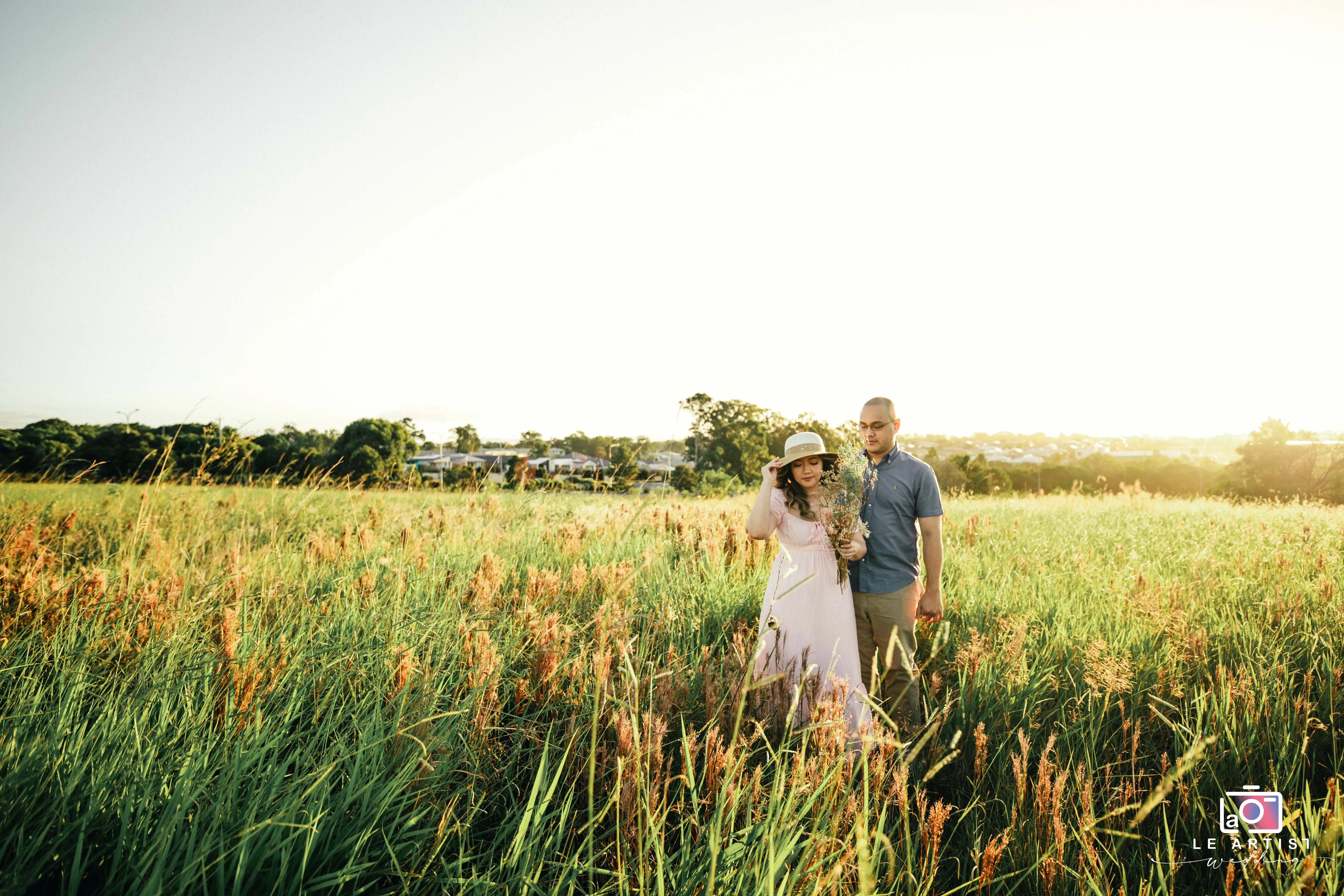 A couple enjoys a peaceful stroll through a sunlit field, capturing a serene and intimate moment amidst nature's beauty. Le Artist Wedding