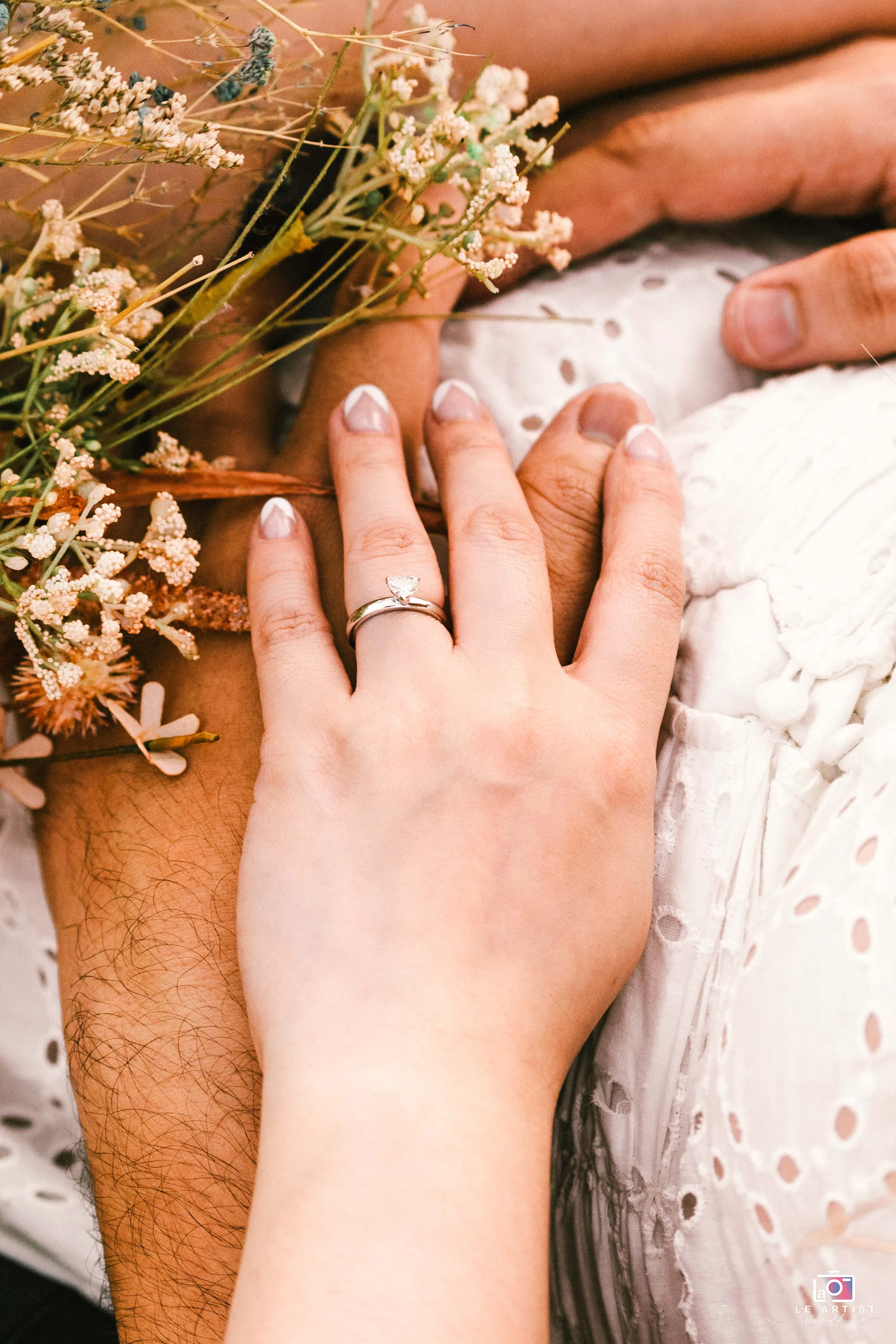 Close-up of intertwined hands adorned with a delicate engagement ring, set against a backdrop of floral lace and dried flowers. Le Artist Wedding