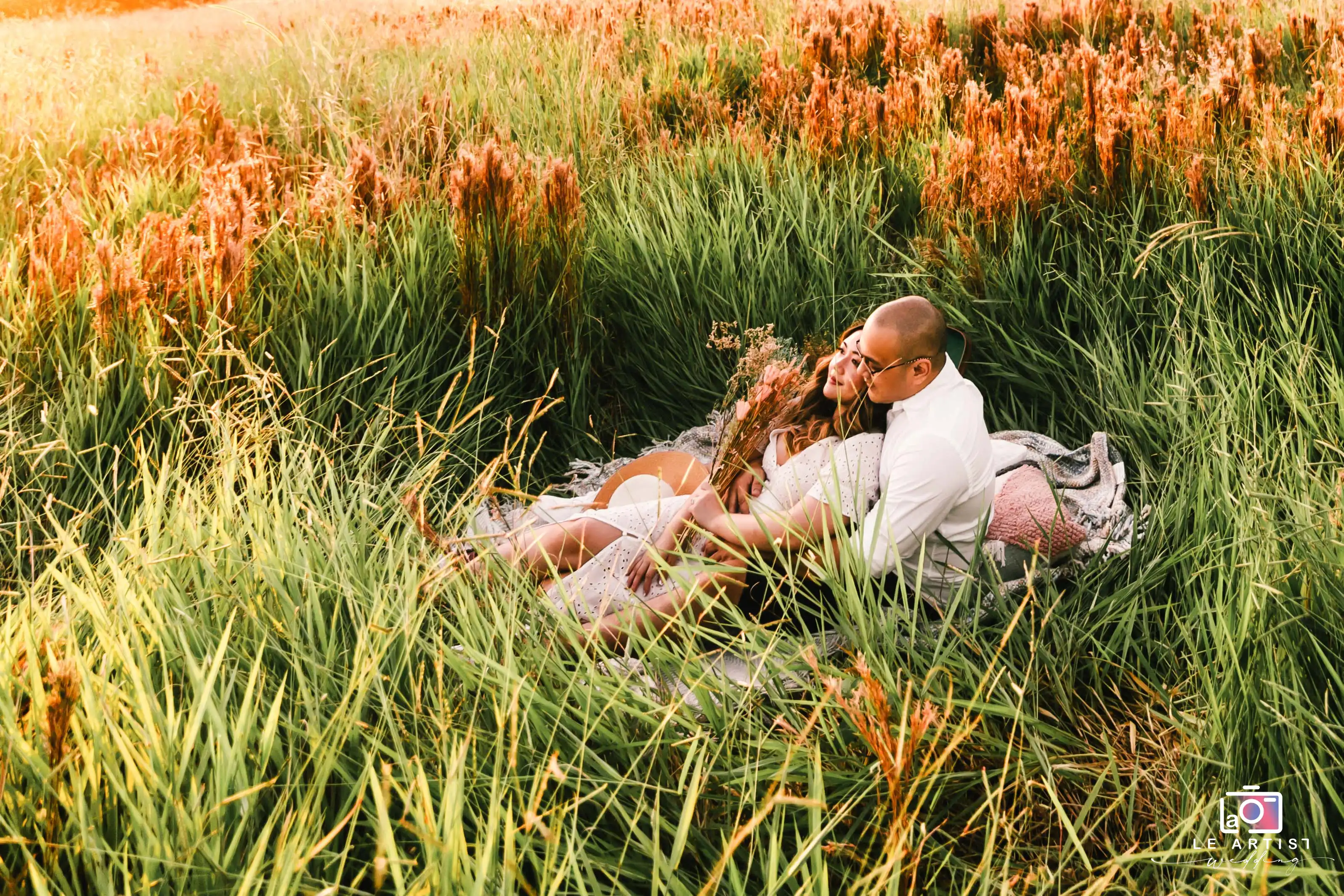 A couple shares an intimate moment, nestled in a lush field at sunset, surrounded by tall grass and warm light. Le Artist Wedding