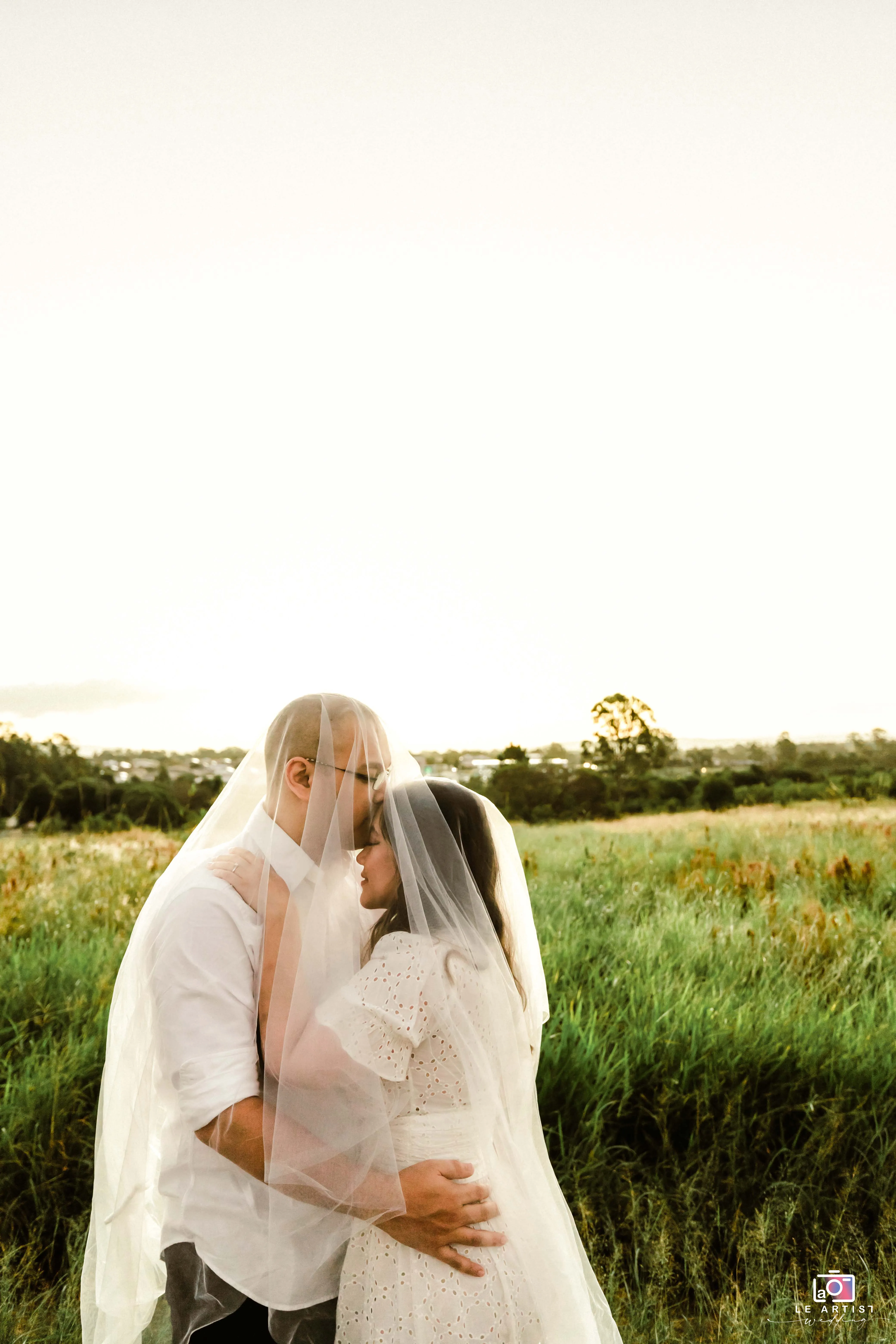 The couple shares an intimate moment under the veil amidst a serene outdoor setting, embodying the romantic essence of the day. Le Artist Wedding