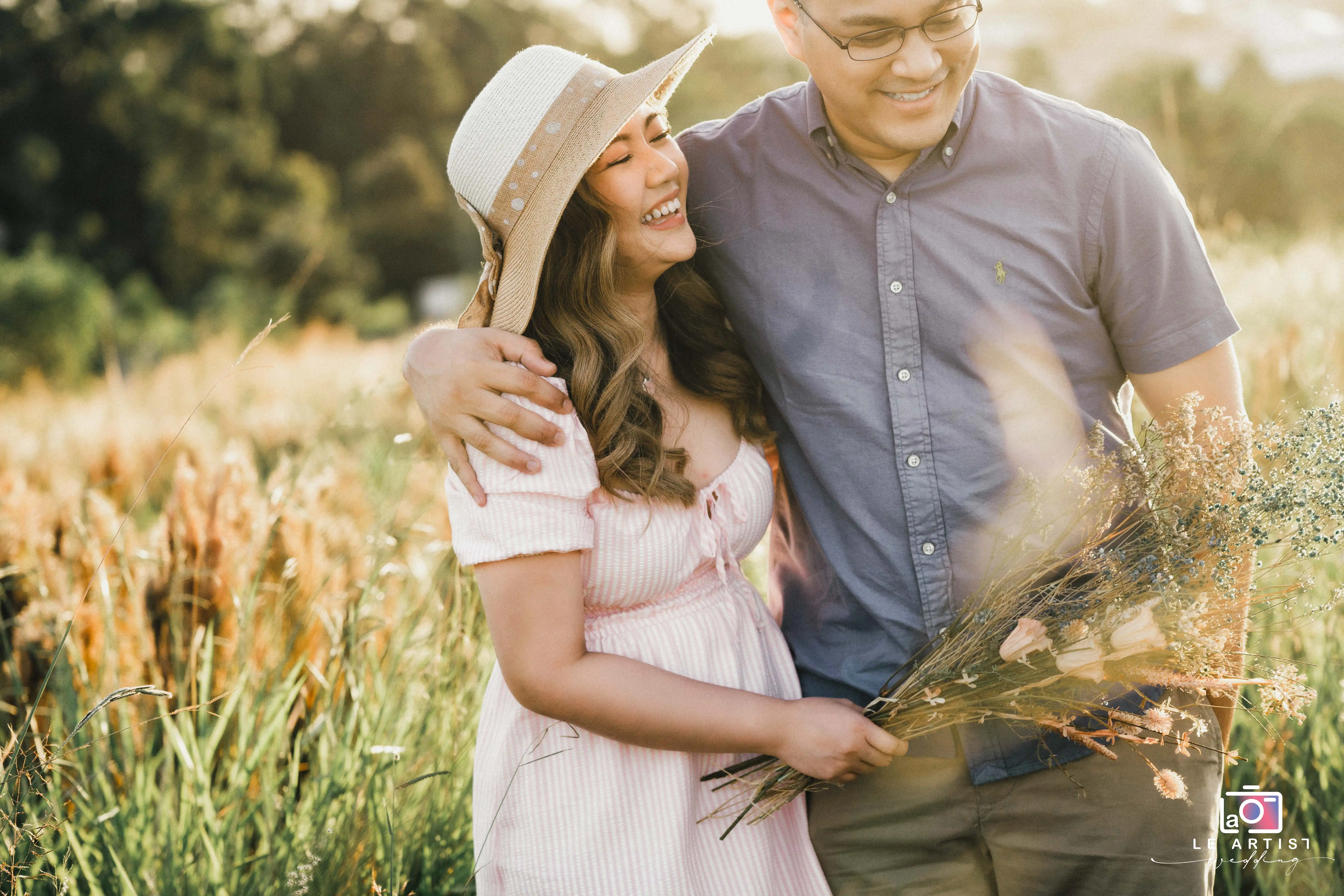 A joyful couple embracing in a sunlit field, sharing a moment of laughter and warmth. The bride holds a bouquet of wildflowers. Le Artist Wedding