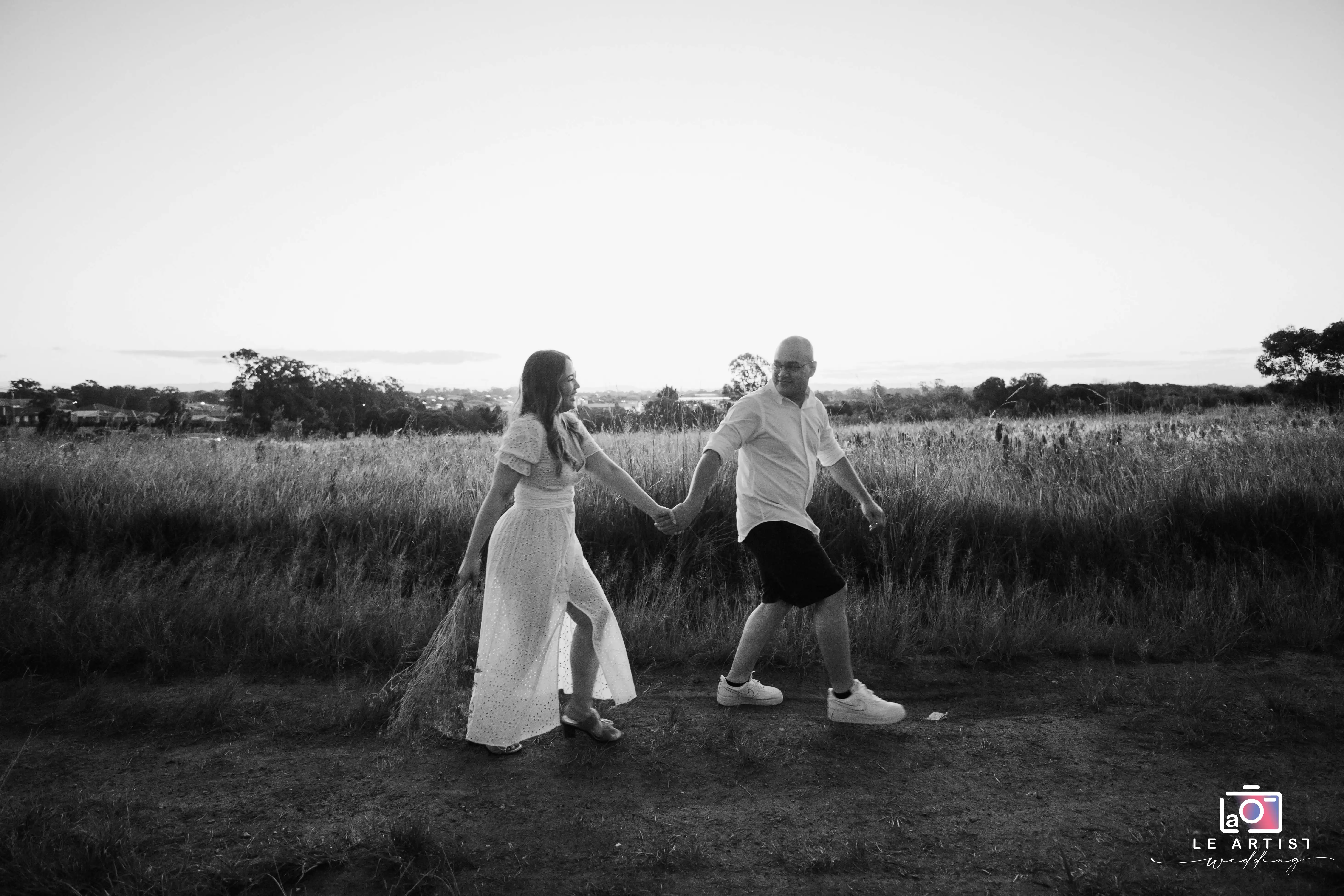 A joyful couple strolls hand in hand through a serene field, capturing a candid moment of love and happiness. Le Artist Wedding