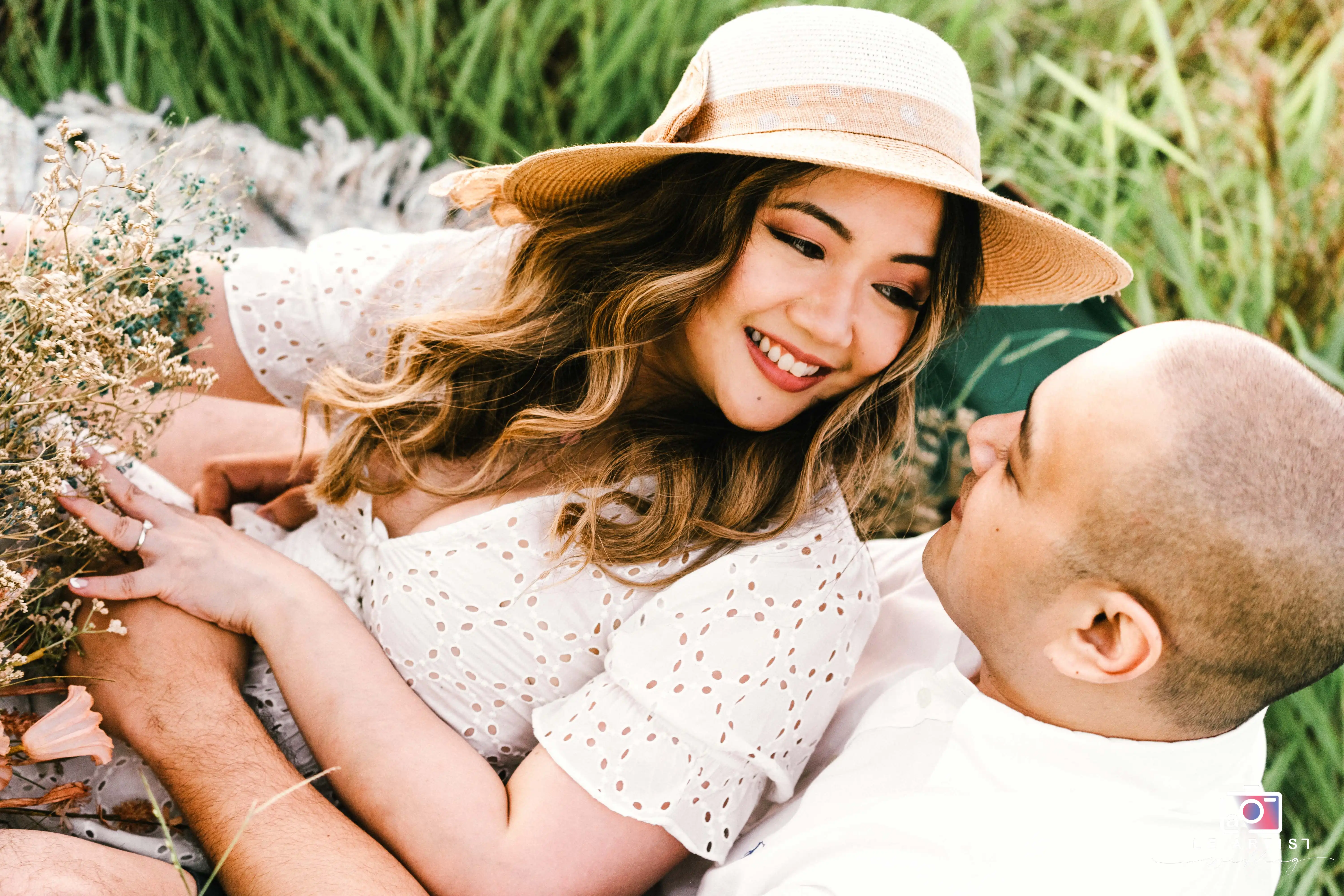 A couple shares a tender moment outdoors, surrounded by greenery, as the bride-to-be lovingly gazes wearing a sun hat. Le Artist Wedding
