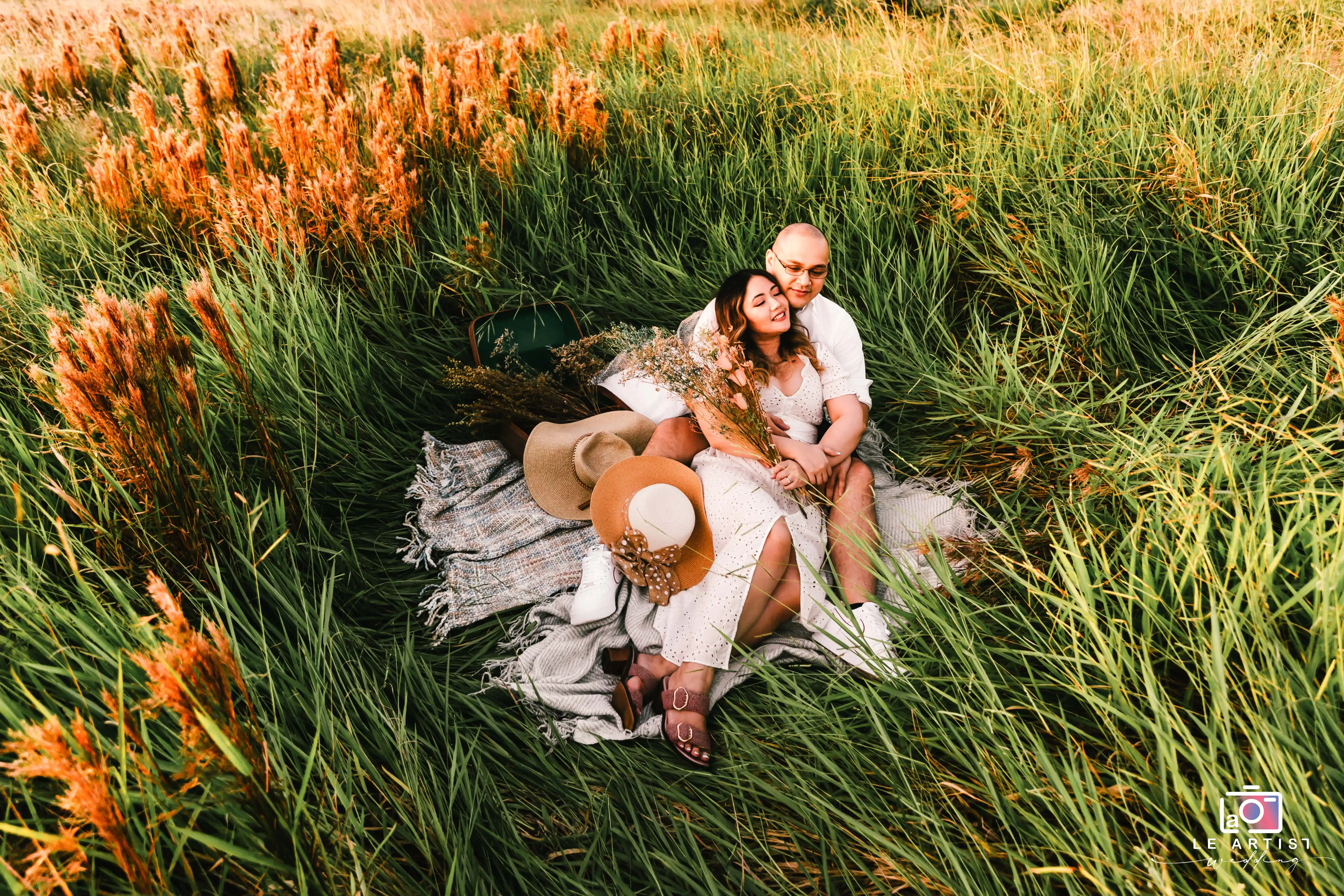 A couple enjoys a romantic moment seated in a lush field, surrounded by golden stalks and greenery, symbolizing tranquility and love. Le Artist Wedding