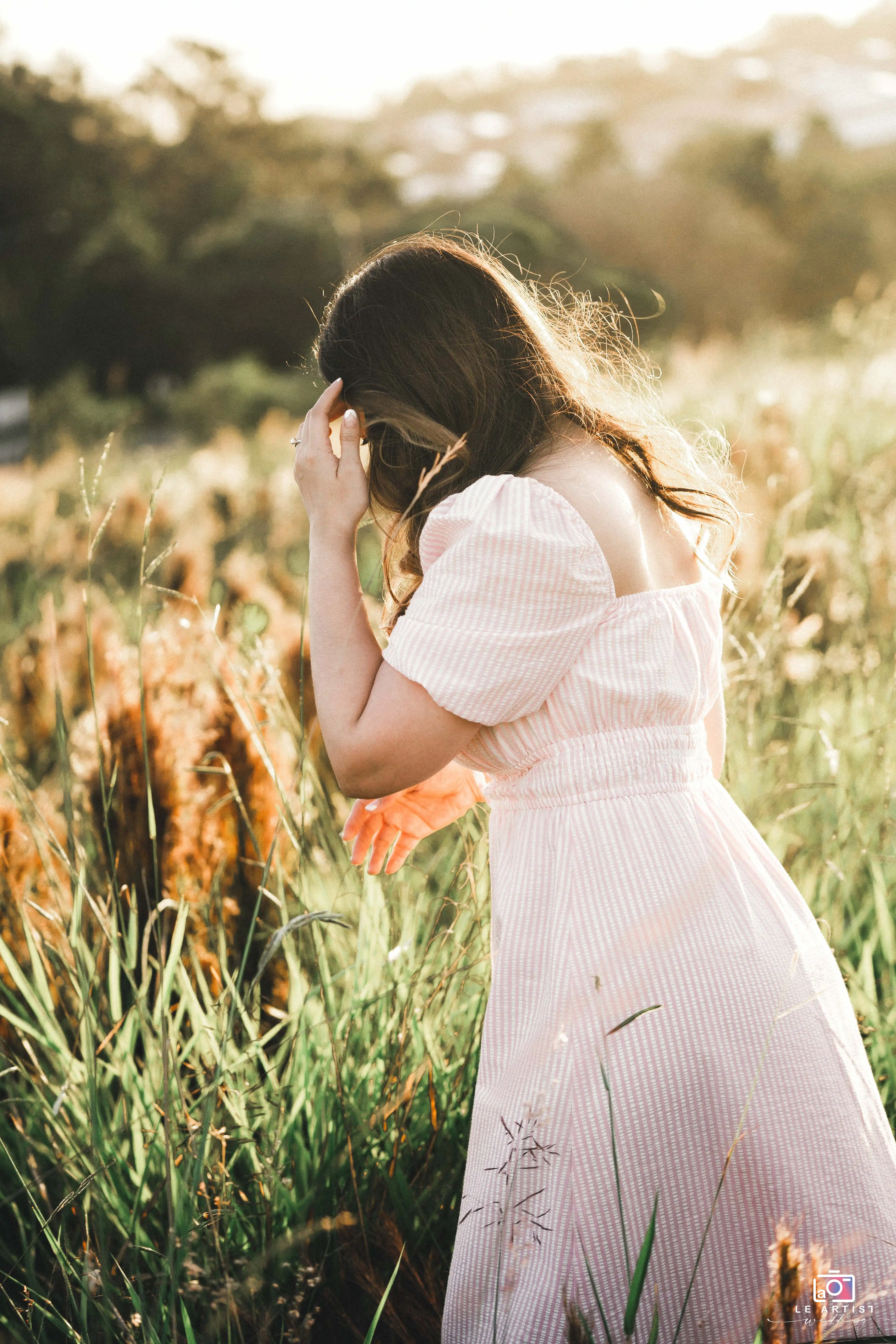 A serene moment captured as a woman in a light pink dress stands thoughtfully in a sunlit field, surrounded by tall grasses. Le Artist Wedding