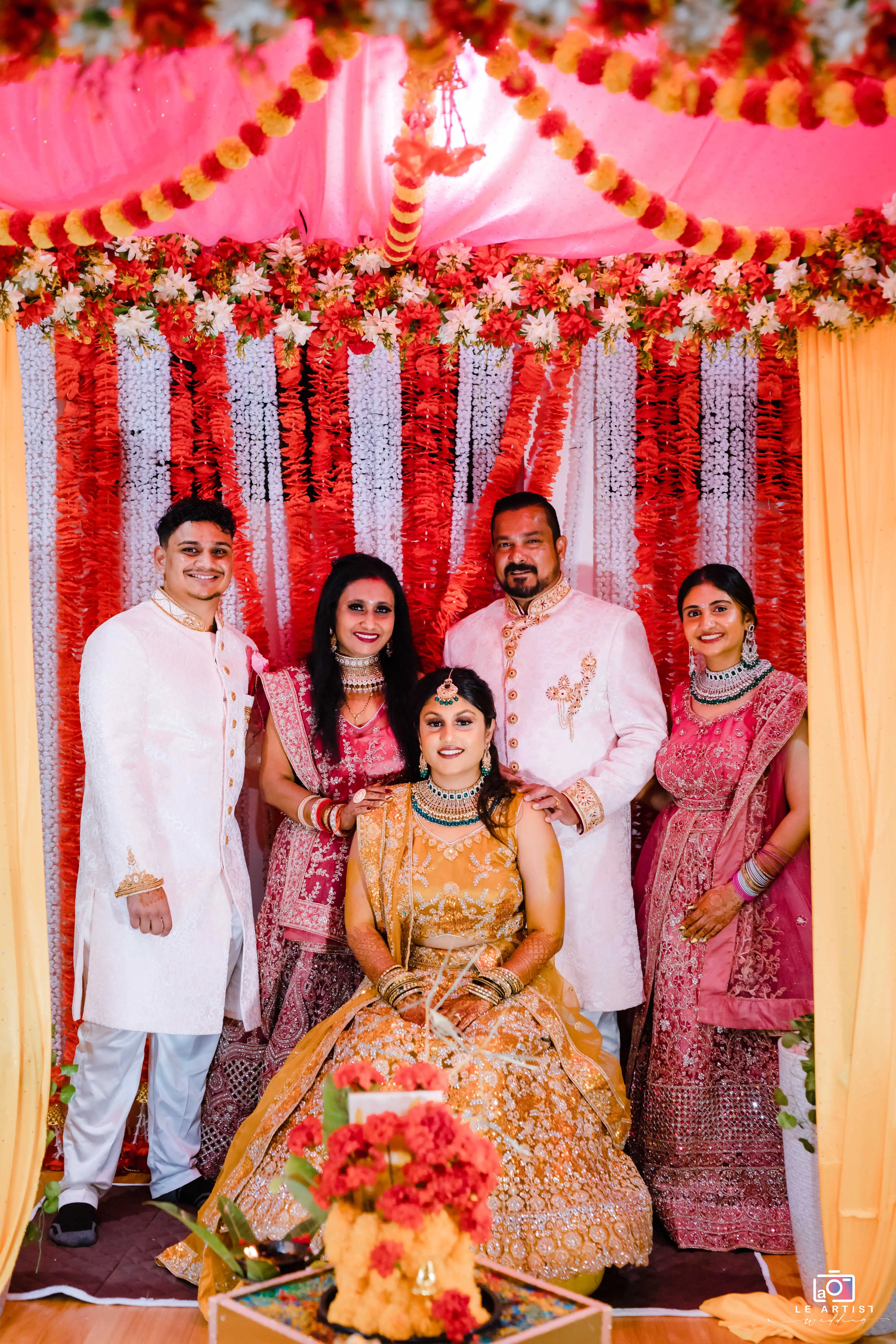 The bridal party poses together under vibrant floral decorations, showcasing traditional attire and joyful expressions. Le Artist Wedding