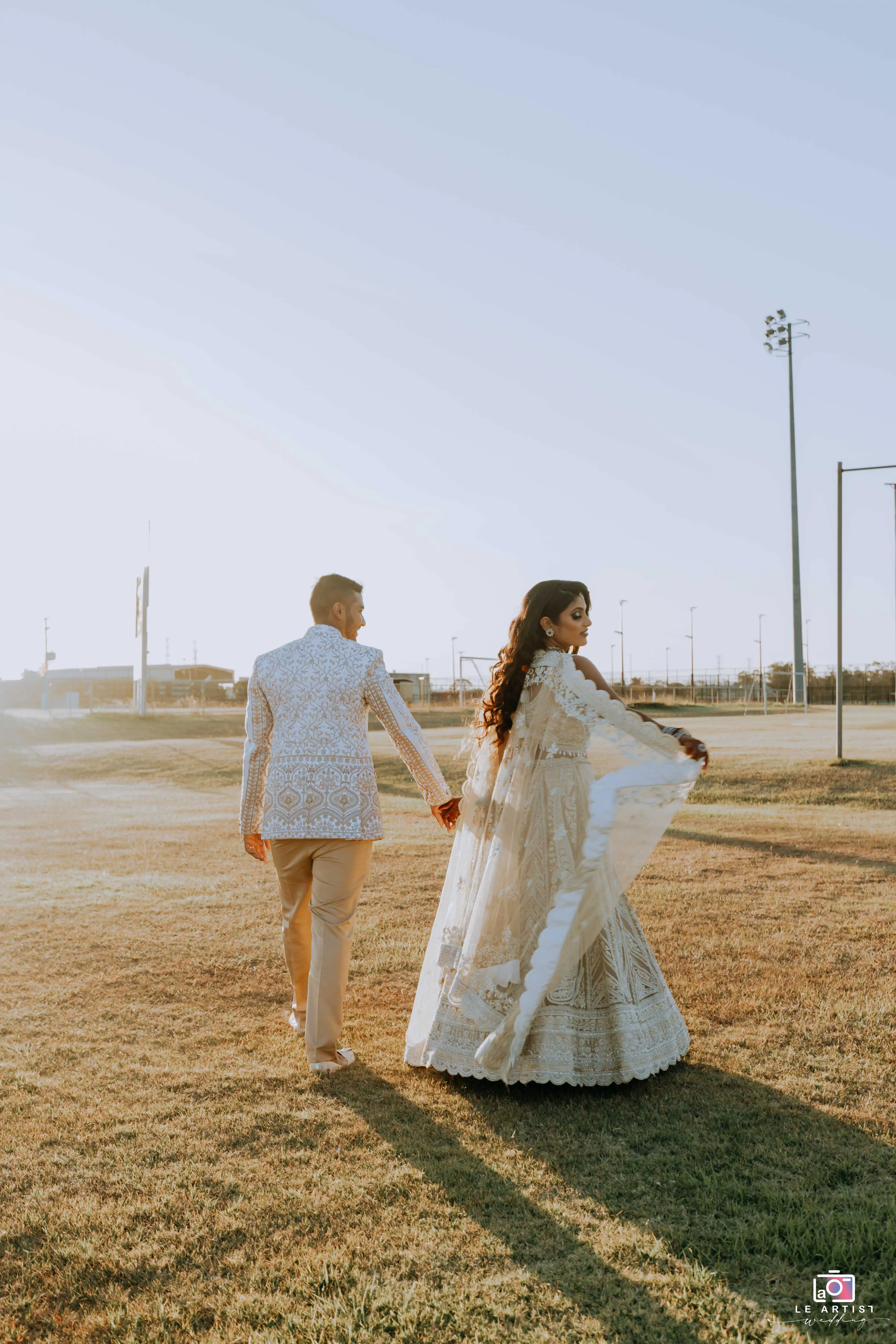 A romantic moment as the couple walks hand in hand in a sunlit field, capturing their love and elegance. Le Artist Wedding