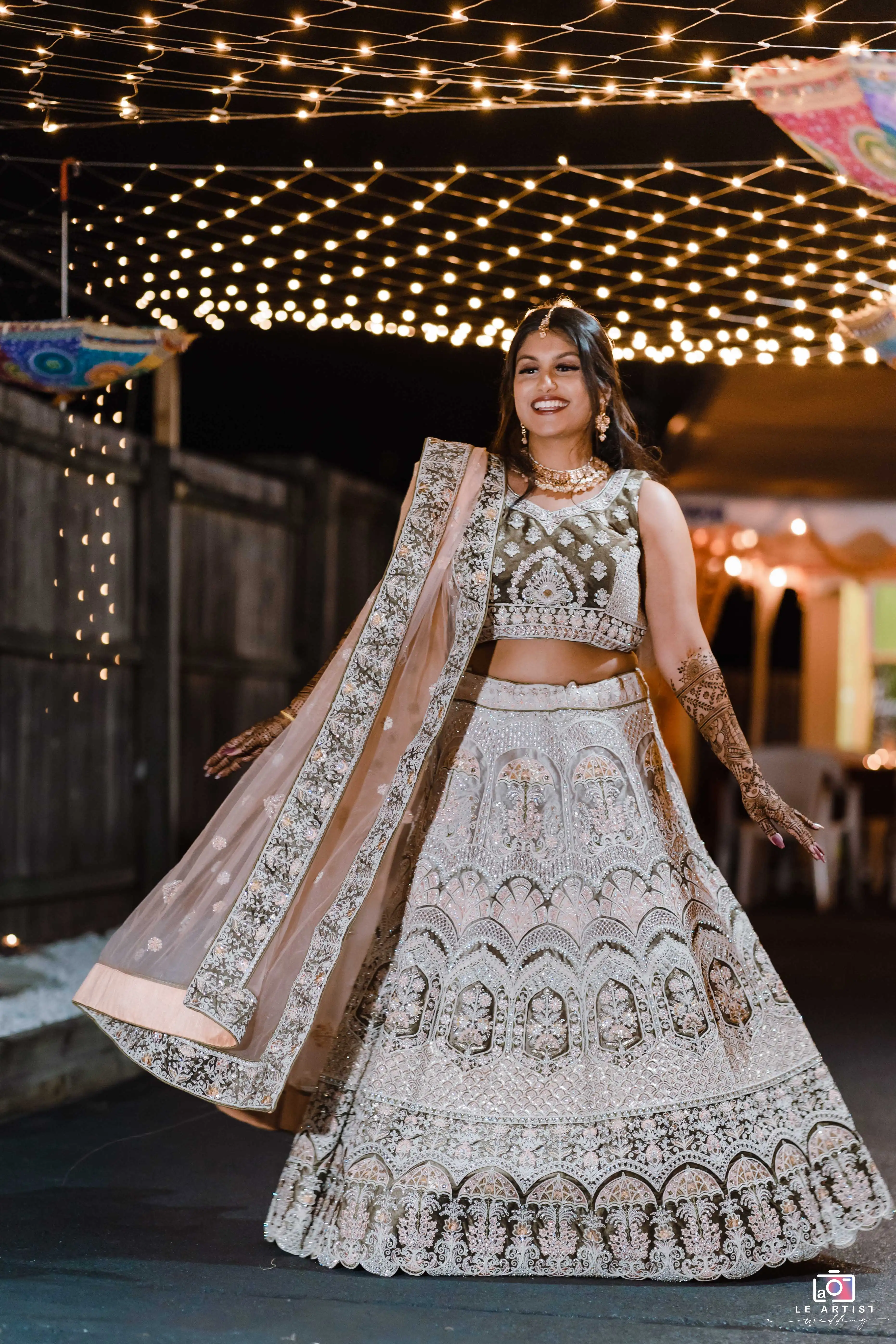 The bride joyfully dances under twinkling lights at the wedding reception, showcasing her intricate attire and vibrant smile. Le Artist Wedding