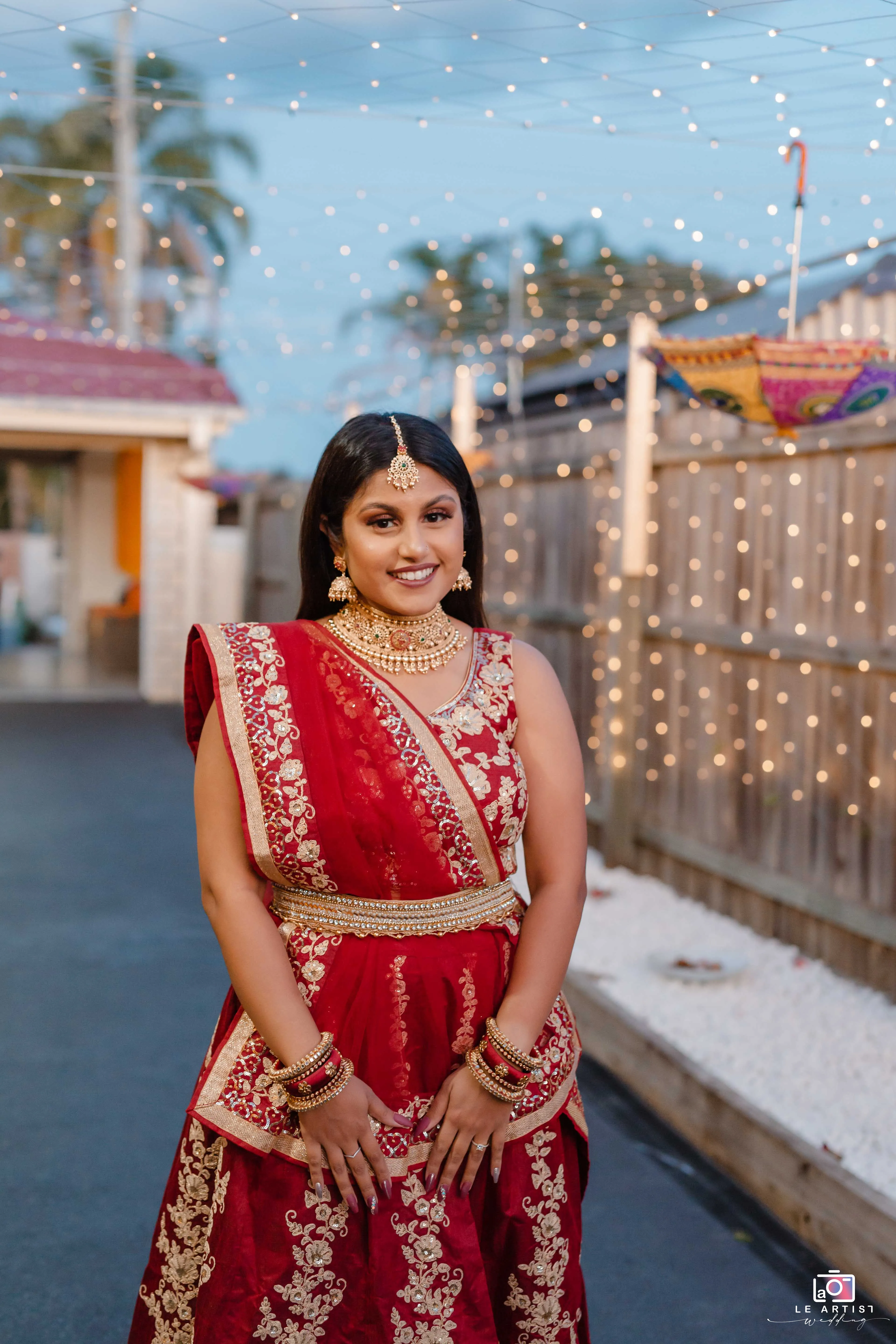 A beautifully dressed bride in a traditional red and gold outfit poses with a warm smile under a canopy of twinkling lights. Le Artist Wedding
