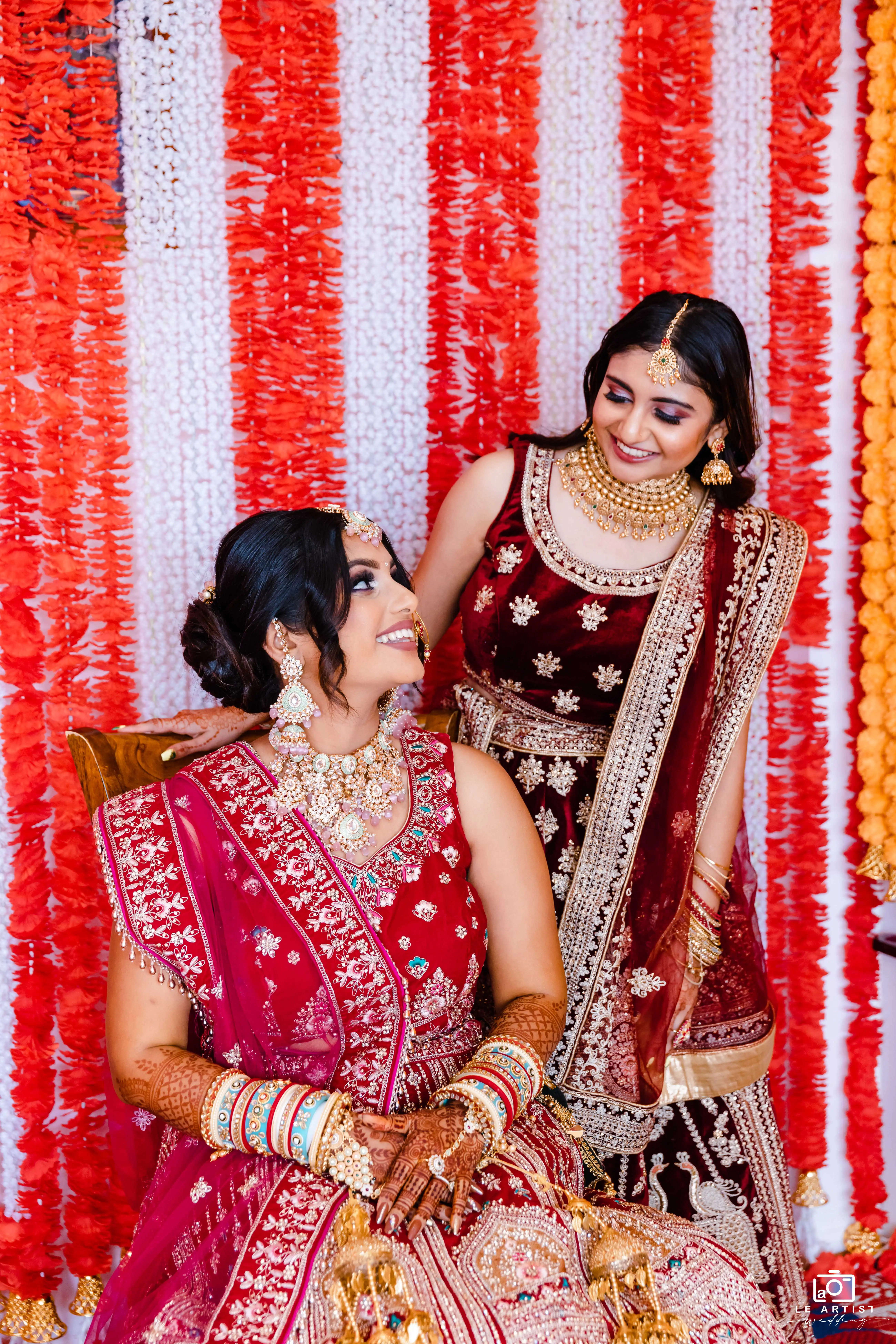 The bride shares a joyful moment with her bridesmaid in front of vibrant floral decorations, showcasing their stunning traditional attire. Le Artist Wedding