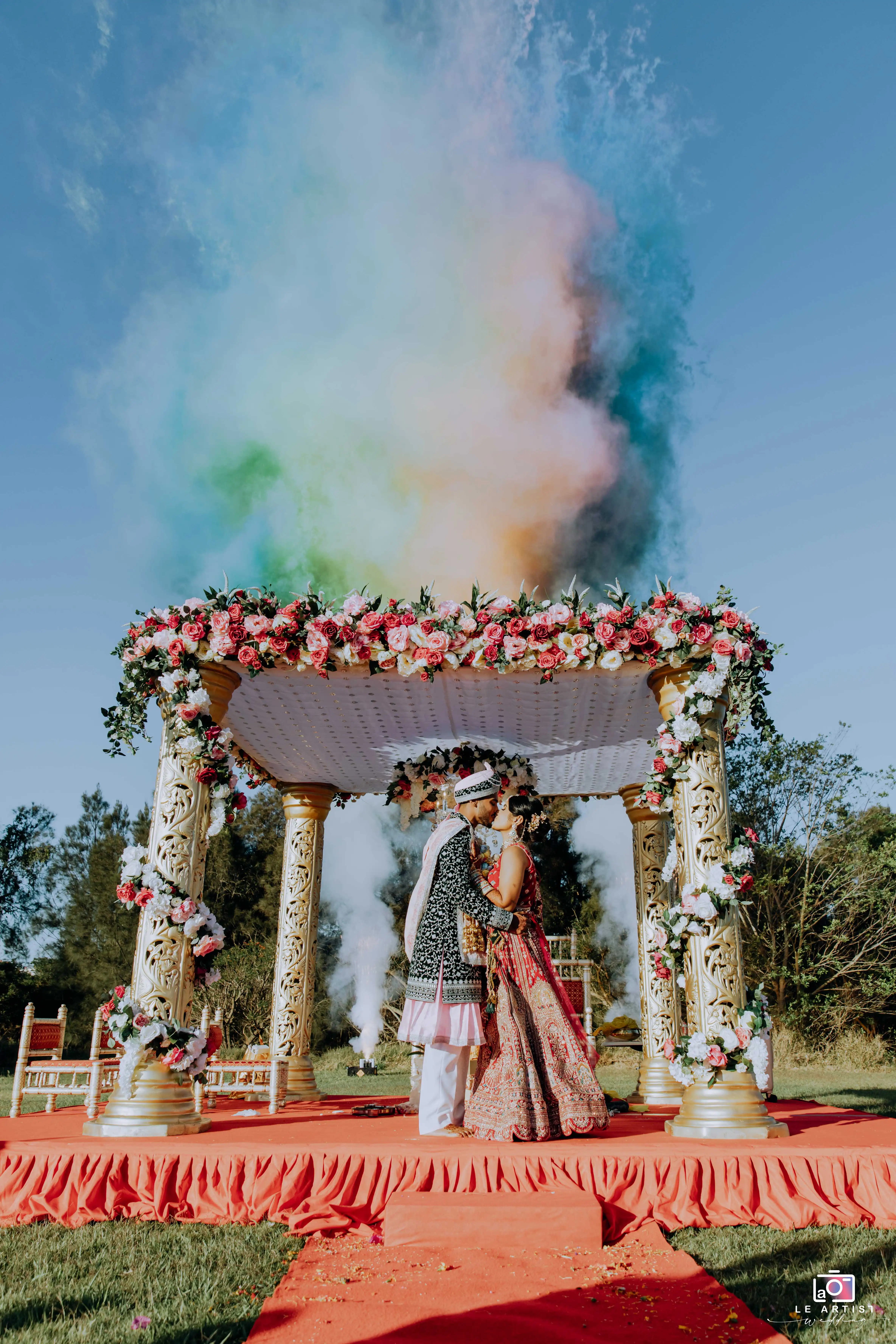 A beautifully adorned mandap serves as the backdrop for the couple's ceremony, with colorful smoke creating a vibrant scene. Le Artist Wedding