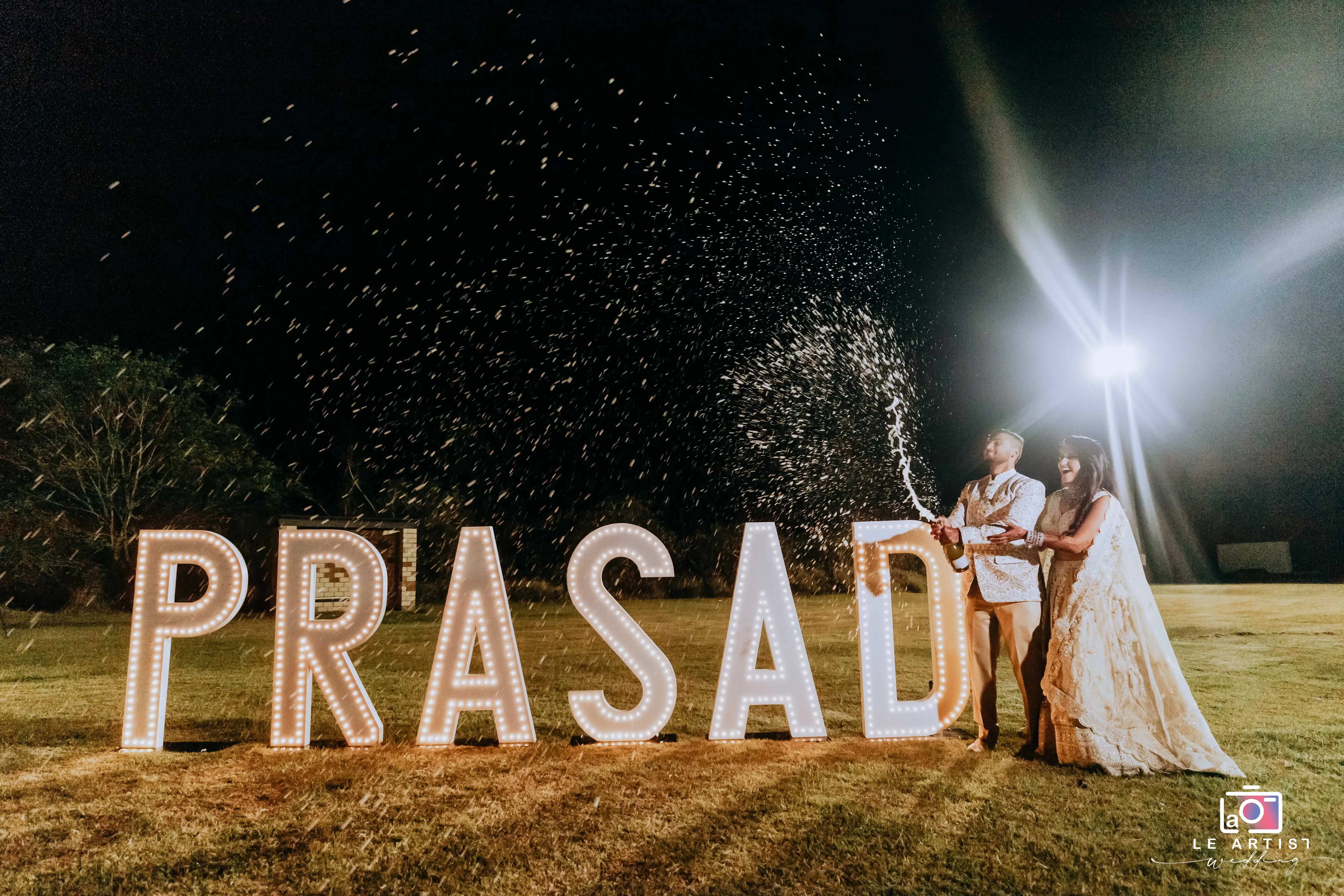 The couple celebrates by popping champagne in front of illuminated letters during the reception. Le Artist Wedding