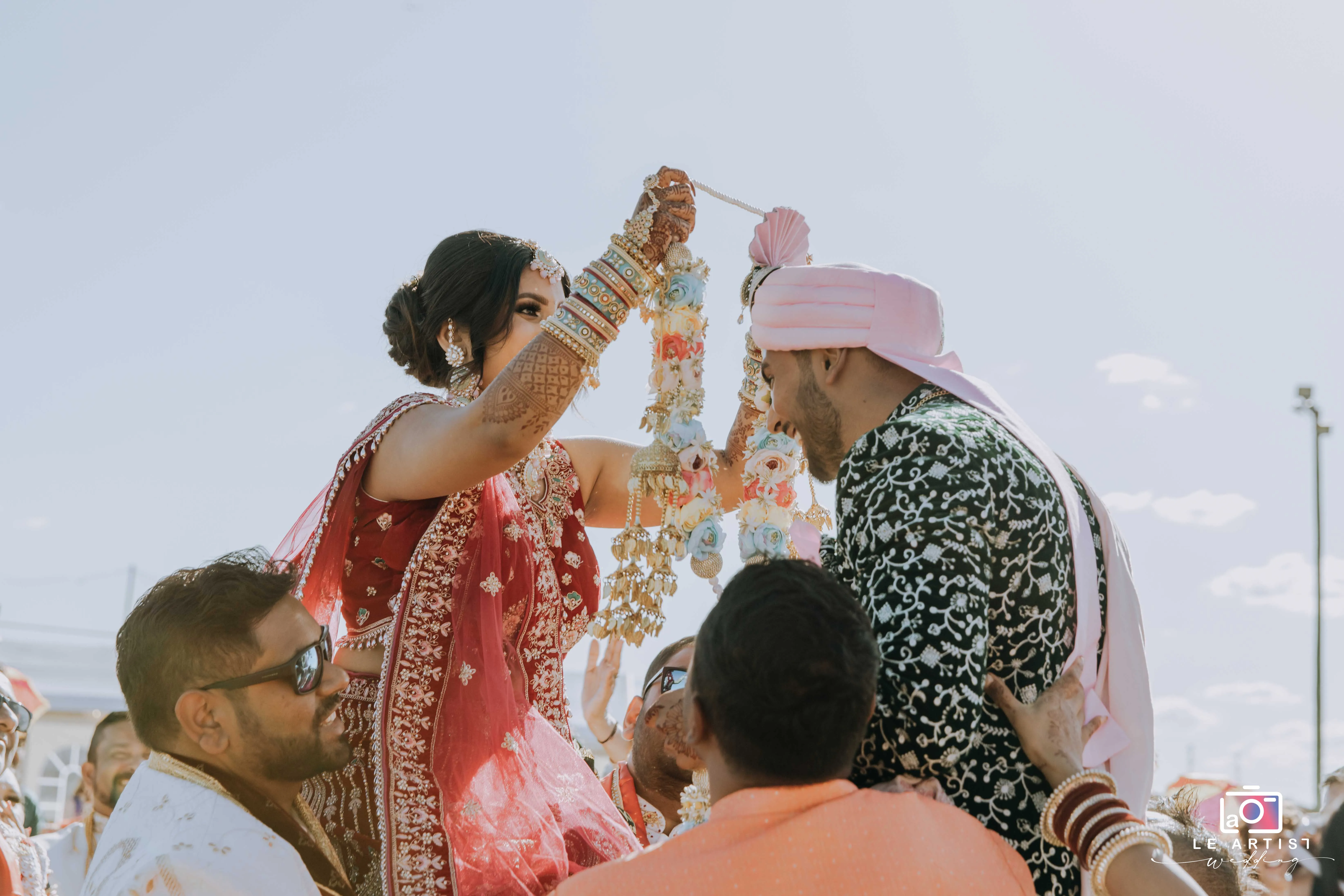 The bride and groom engage in a traditional garland exchange during the wedding ceremony, lifted by friends and family in a joyful celebration. Le Artist Wedding