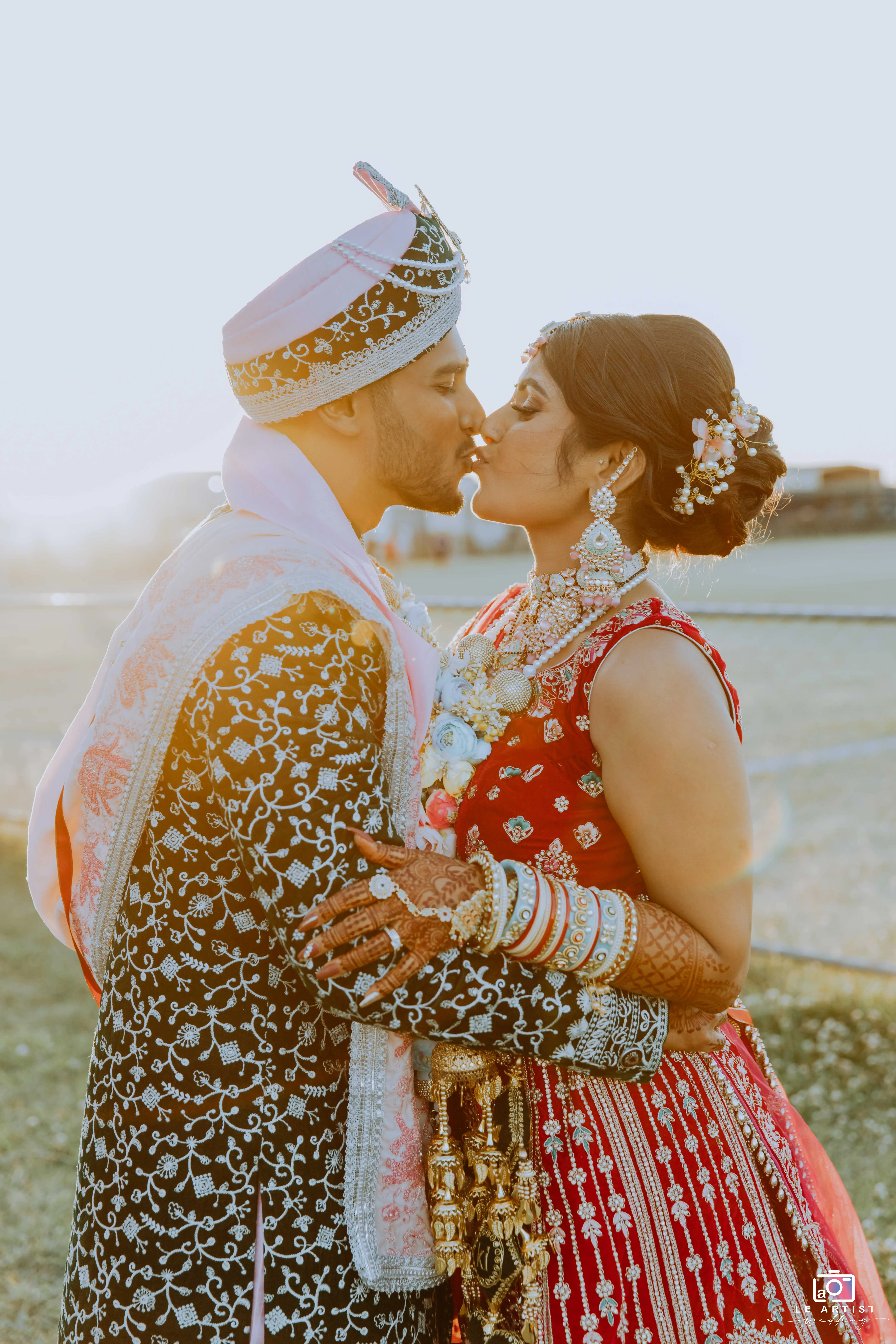 The newlyweds share a romantic kiss, dressed in traditional attire, with the sunset as a beautiful backdrop. Le Artist Wedding