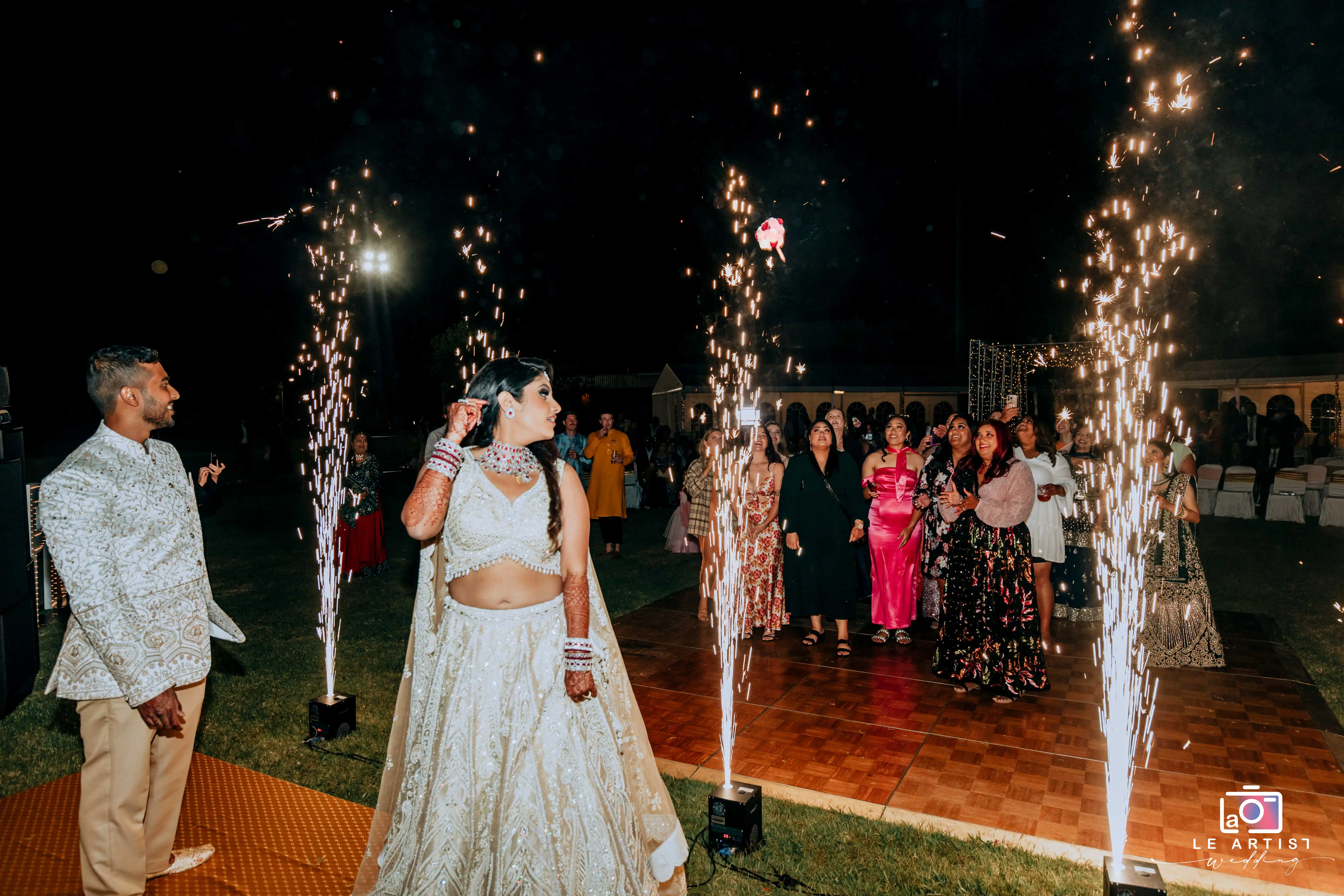 The bride tosses her bouquet amidst sparklers at the reception, while guests eagerly await to catch it. Le Artist Wedding