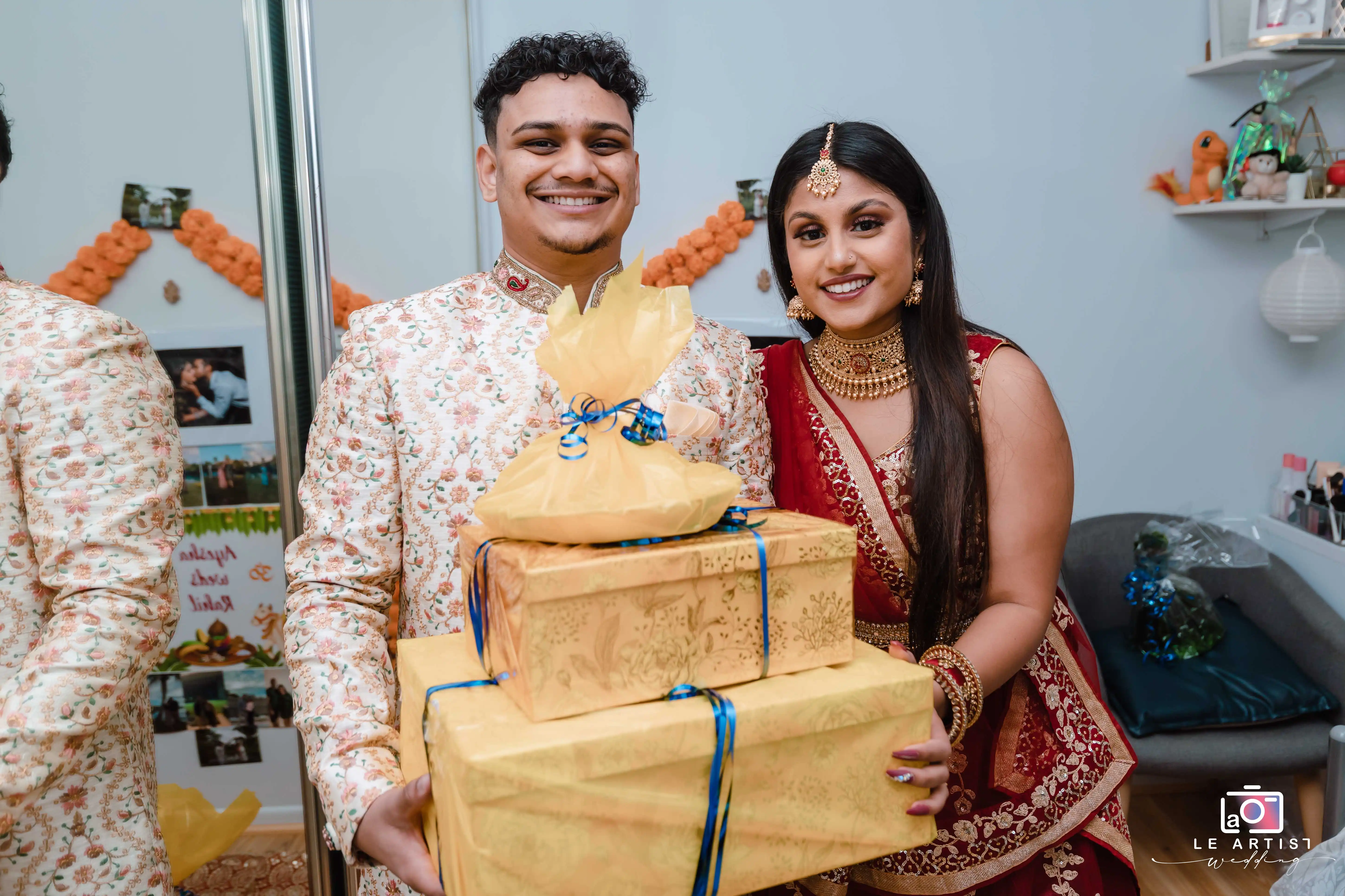 Couple smiling while holding wrapped gifts during the reception. Their attire is beautifully adorned with traditional patterns and jewelry, highlighting the joyous celebration. Le Artist Wedding