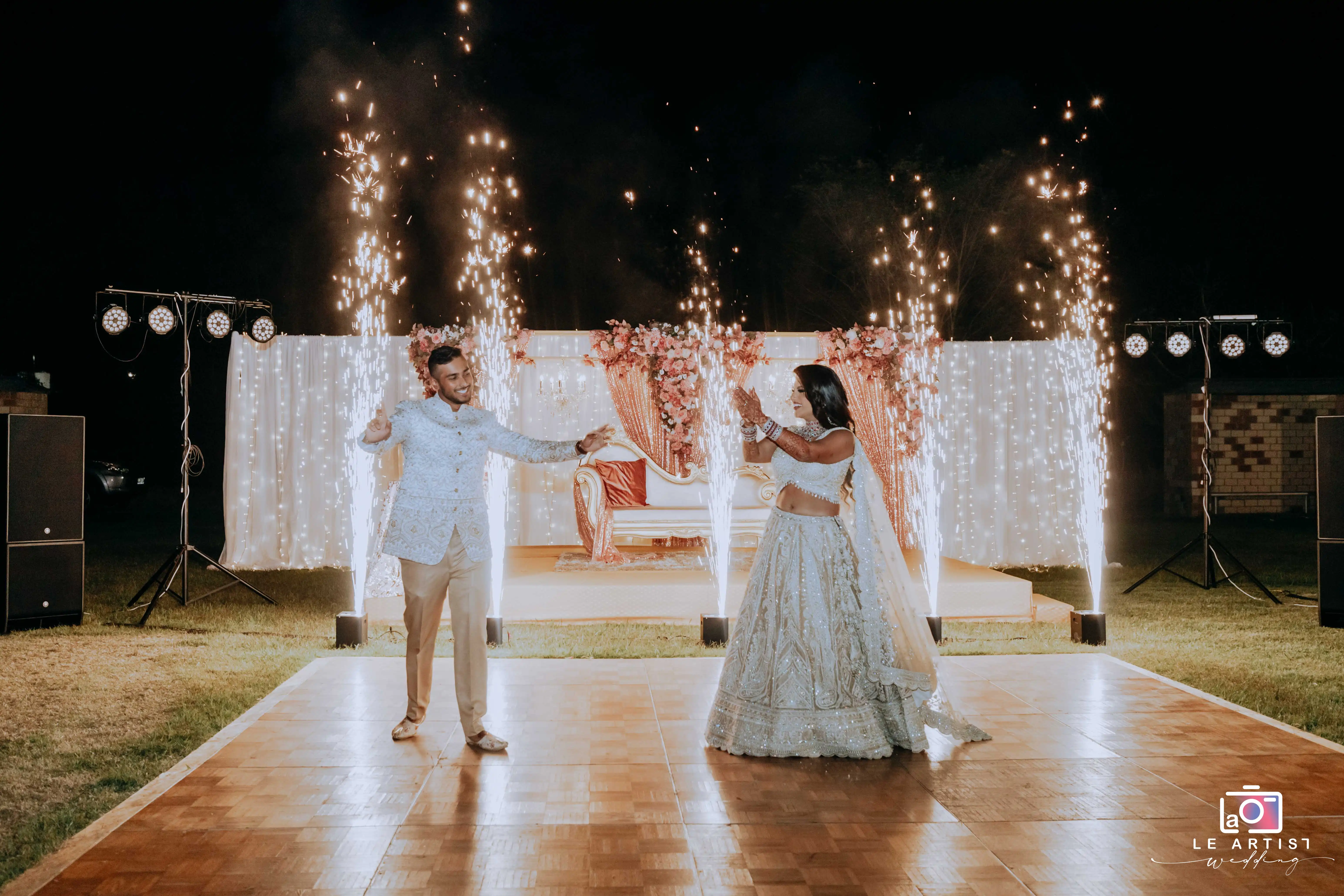 A couple shares a joyful dance surrounded by bright sparklers and an elegantly decorated stage, capturing a festive moment during their wedding reception. Le Artist Wedding