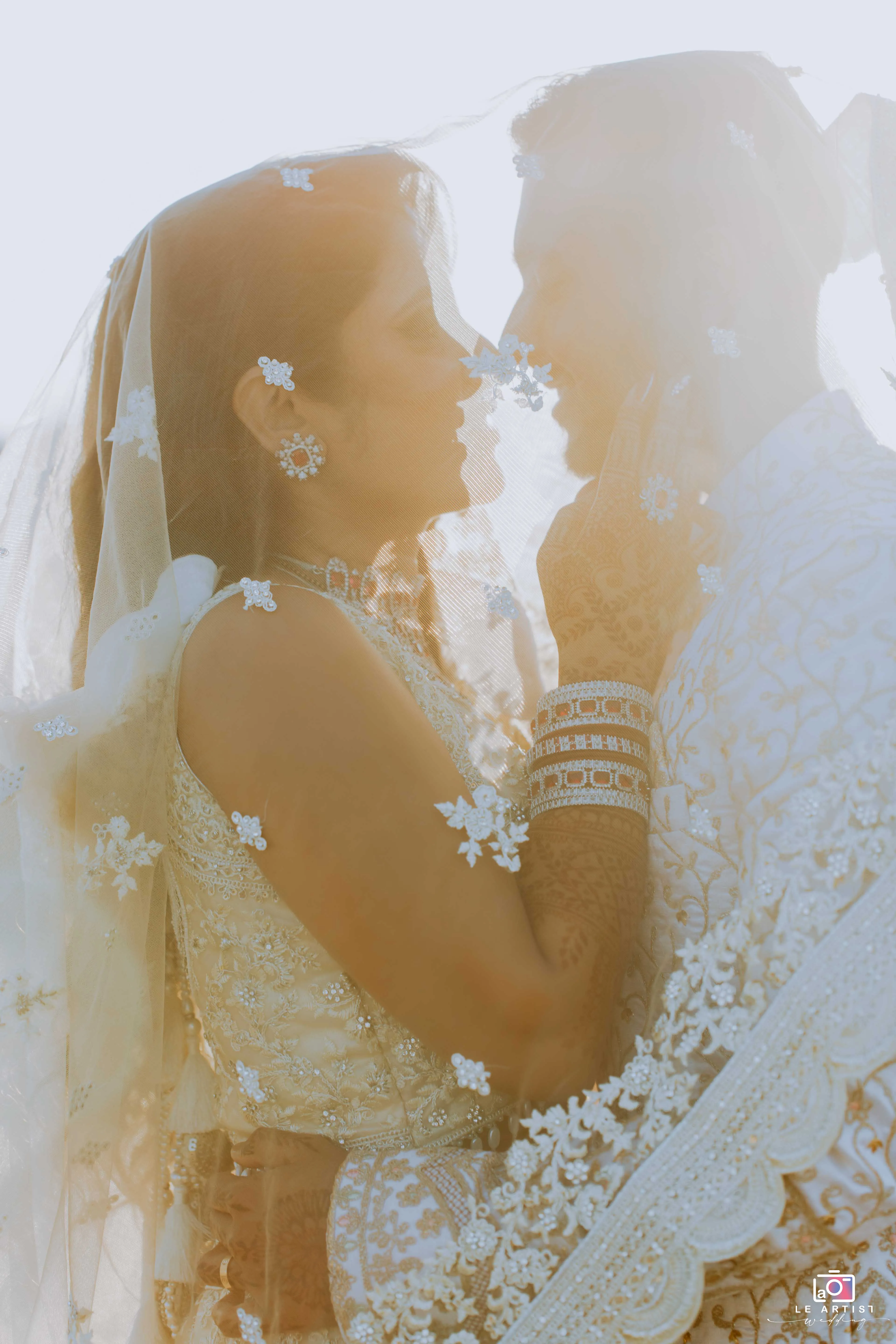 A romantic moment as the couple shares an intimate gaze under a delicate veil, showcasing their intricate attire and jewelry. Le Artist Wedding