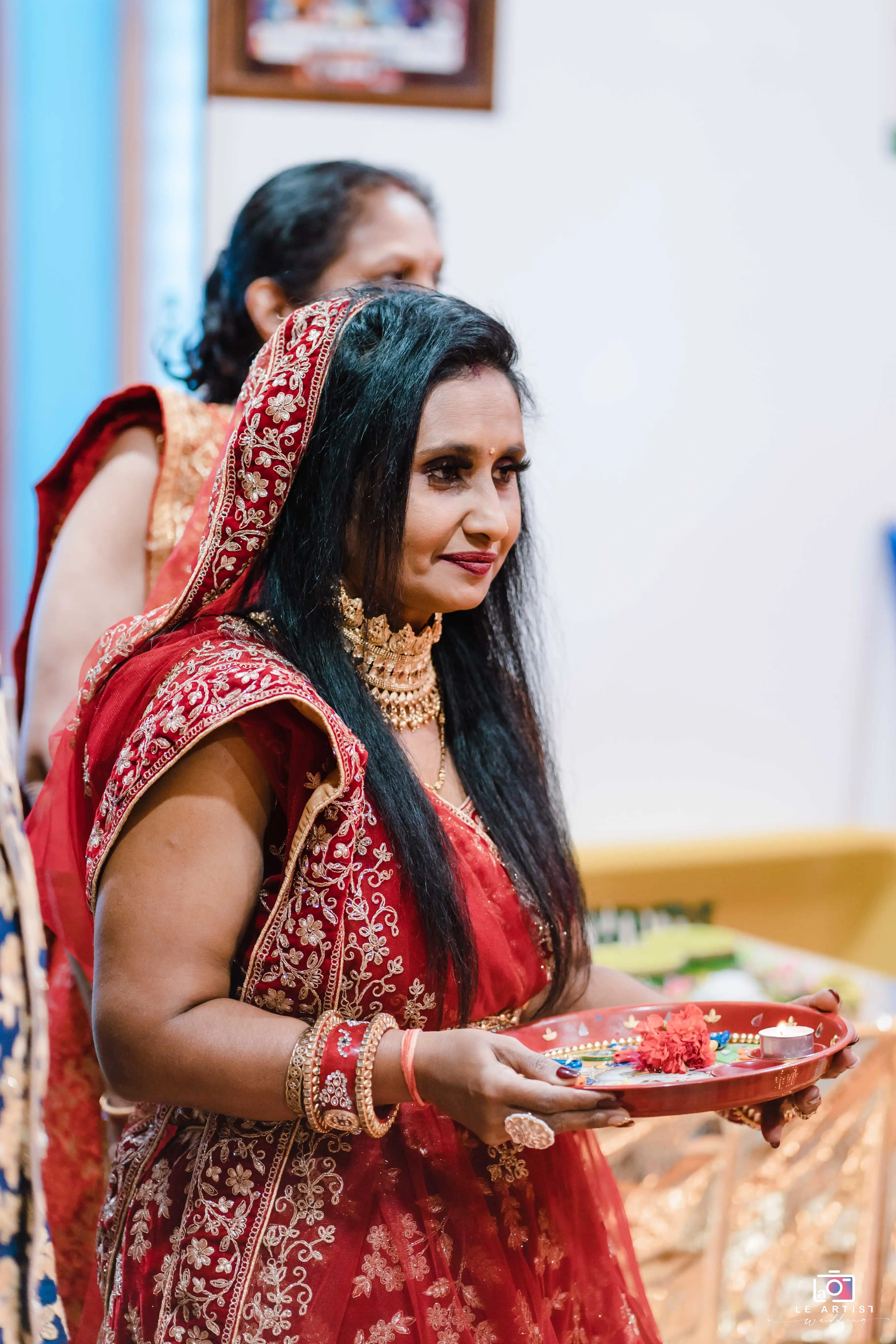 A woman in traditional red attire holds a decorative tray, participating in a cultural marriage ceremony. Le Artist Wedding