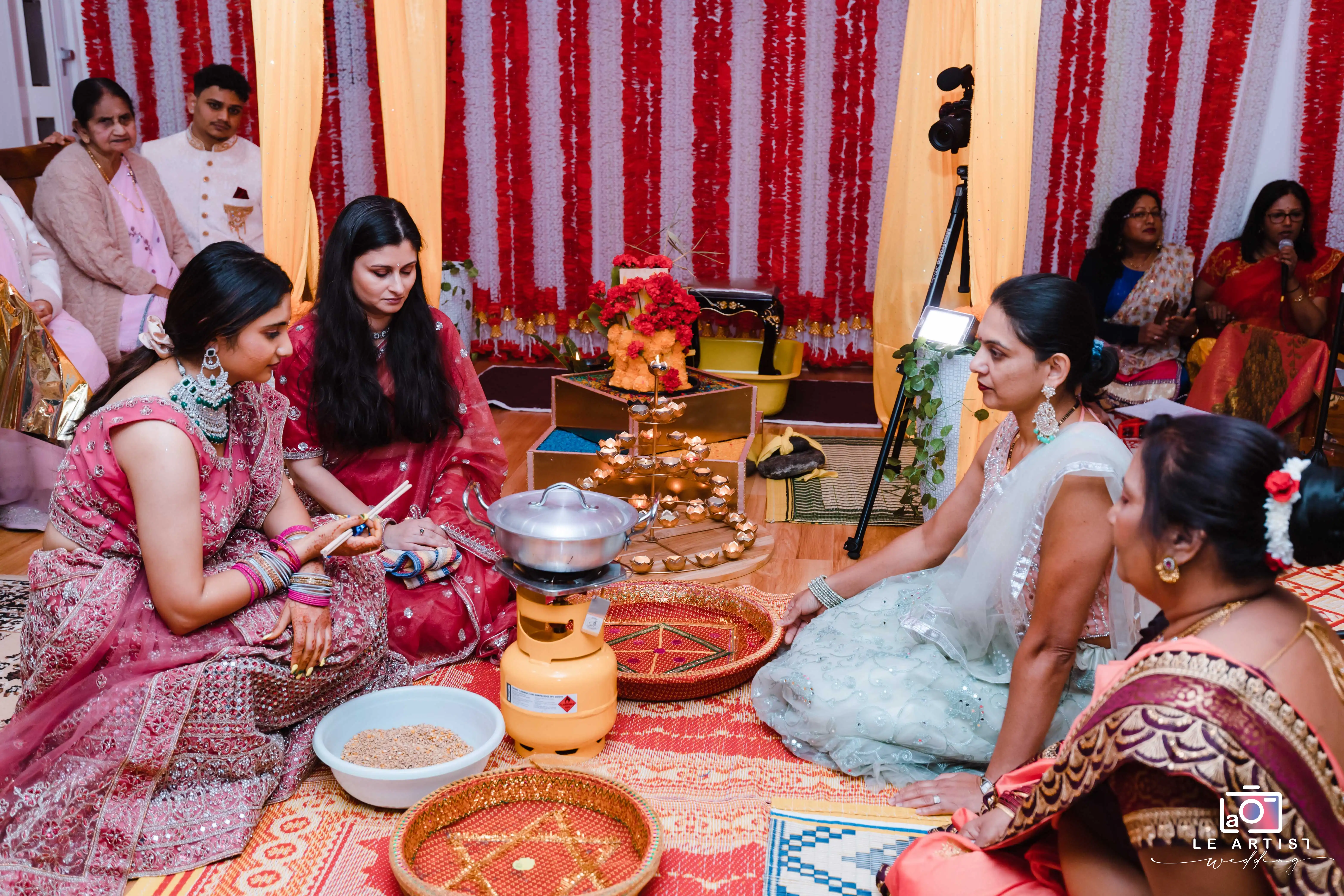 A traditional wedding ritual takes place with family members gathered around ceremonial items, creating an intimate and vibrant setting. Le Artist Wedding