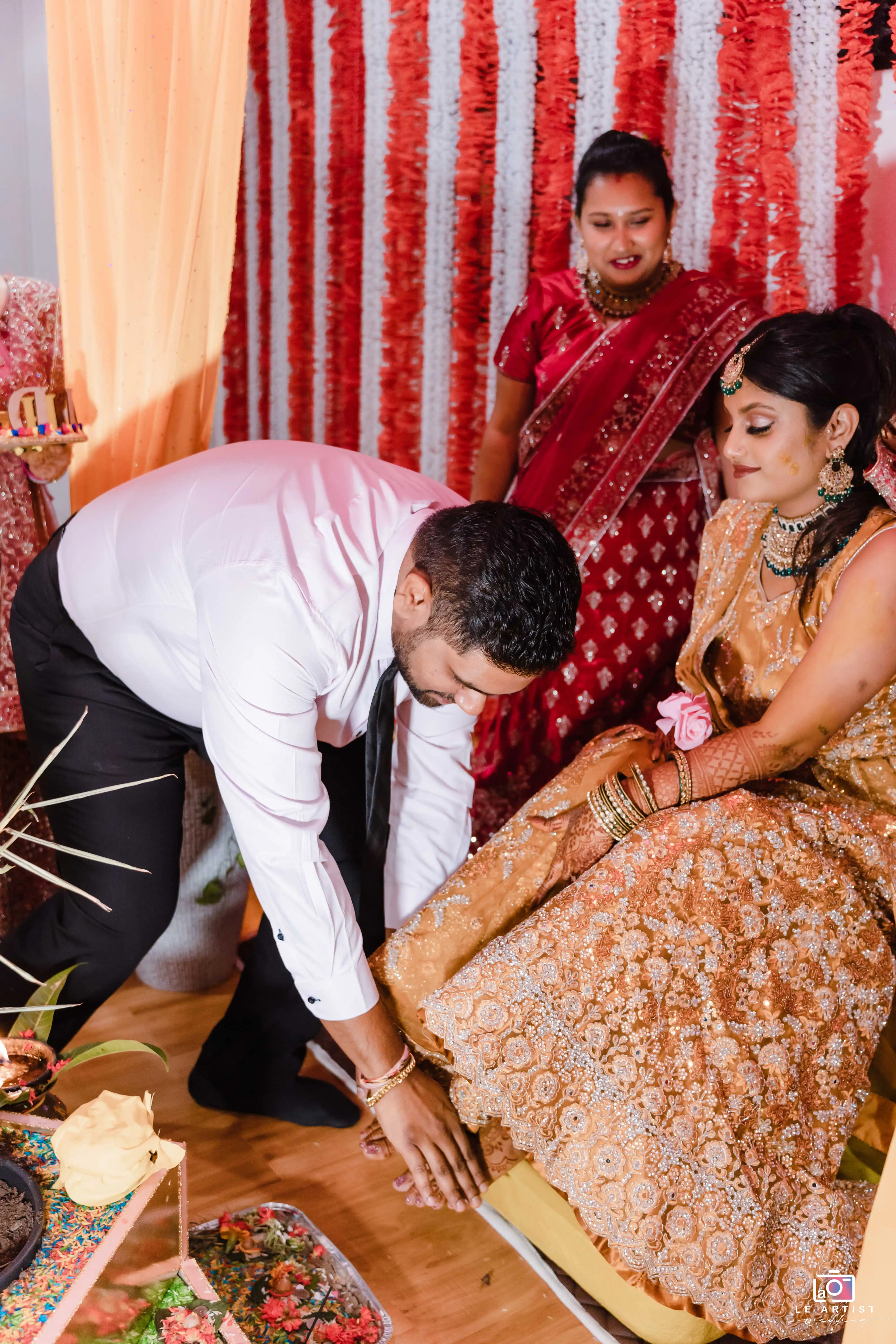 A groom helps the bride during a traditional wedding ceremony ritual, surrounded by vibrant decorations. Le Artist Wedding