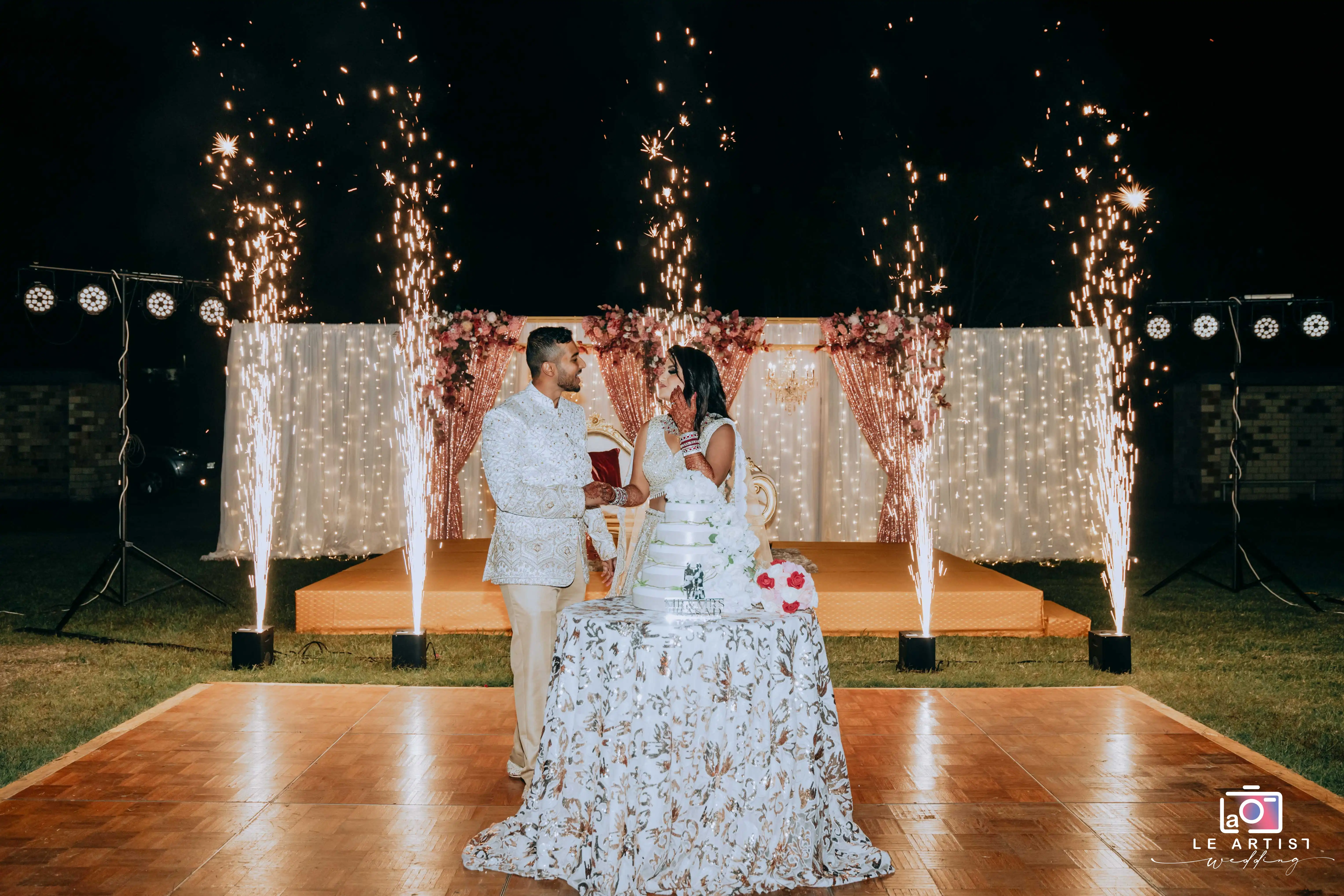 The couple shares a moment before cutting their wedding cake, surrounded by spectacular sparklers and elegant decor, creating a festive reception atmosphere. Le Artist Wedding