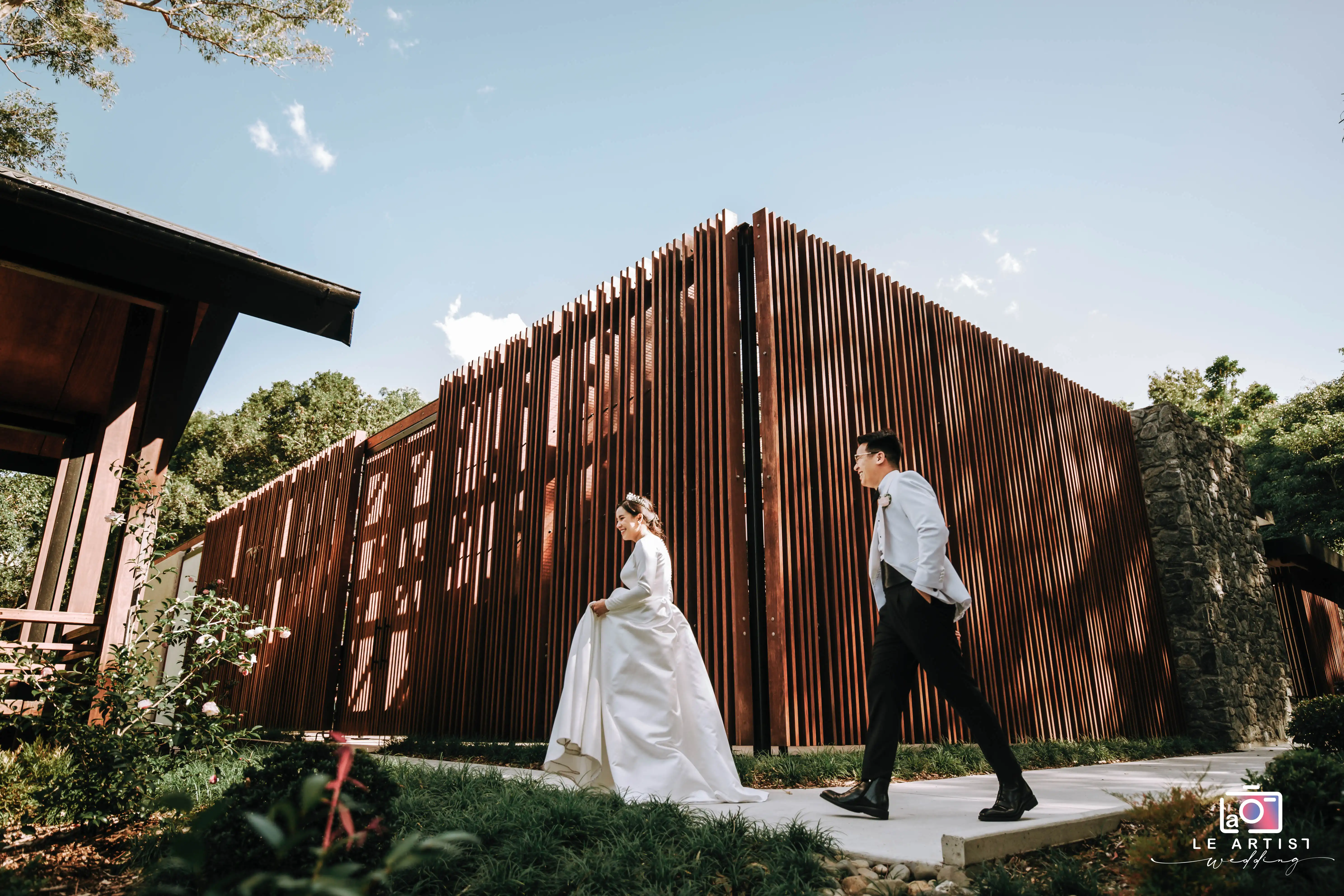 The bride and groom stroll through a serene outdoor setting, capturing a moment of elegance and joy during their wedding photoshoot. Le Artist Wedding