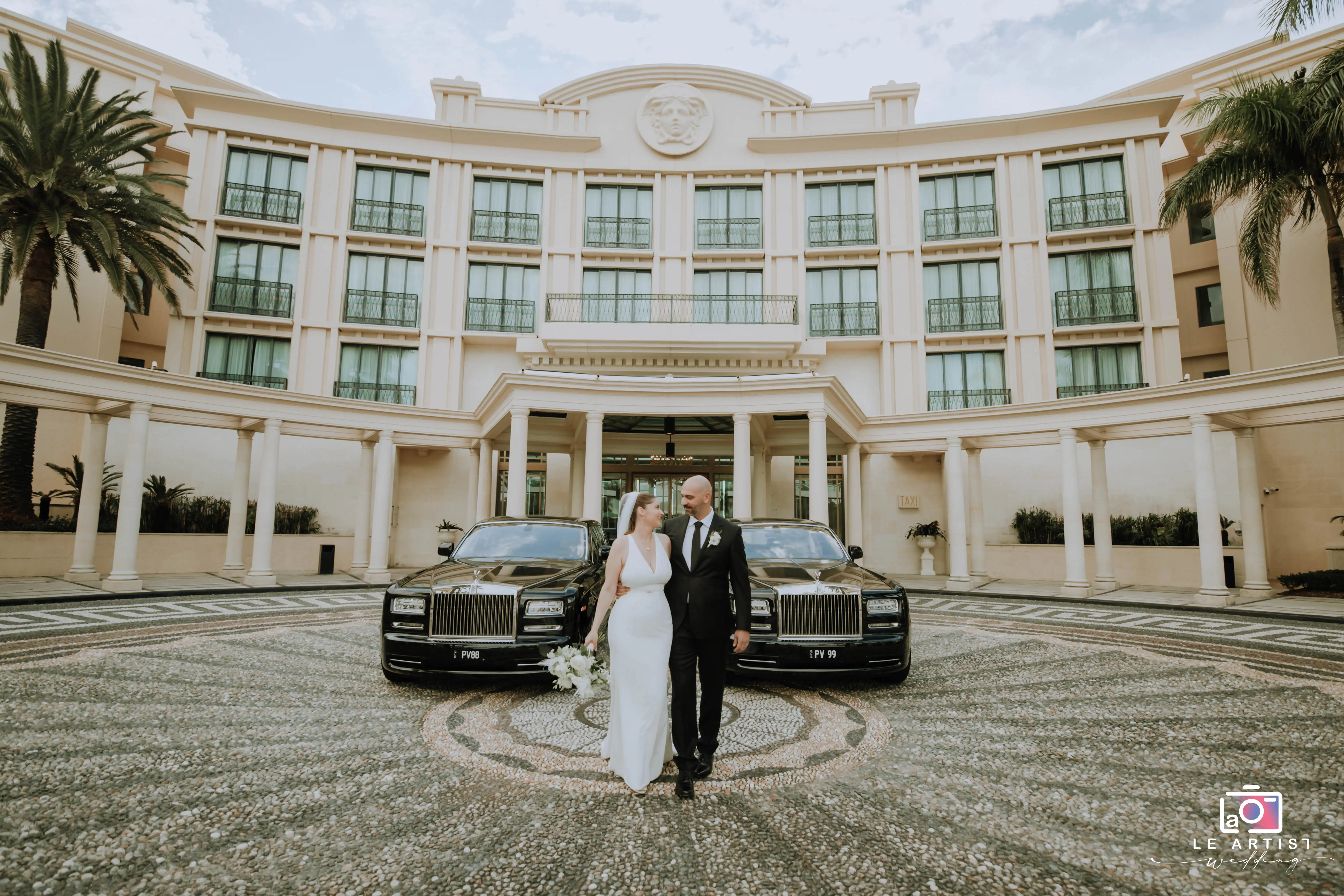The bride and groom elegantly pose in front of a grand building, flanked by luxury cars, capturing a moment of sophisticated celebration. Le Artist Wedding