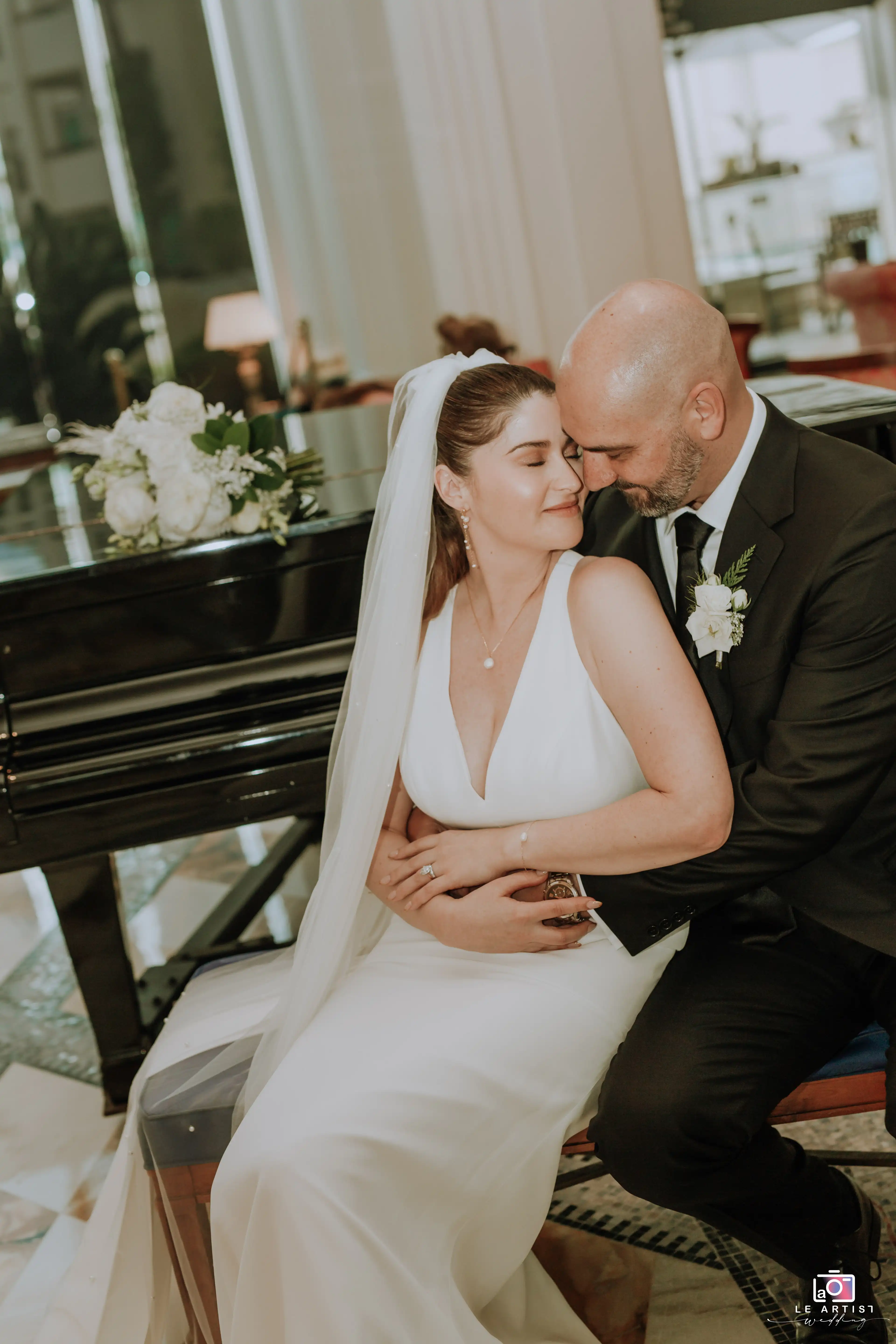 The bride and groom share a tender moment by the piano, showcasing their love and elegance on their special day. Le Artist Wedding