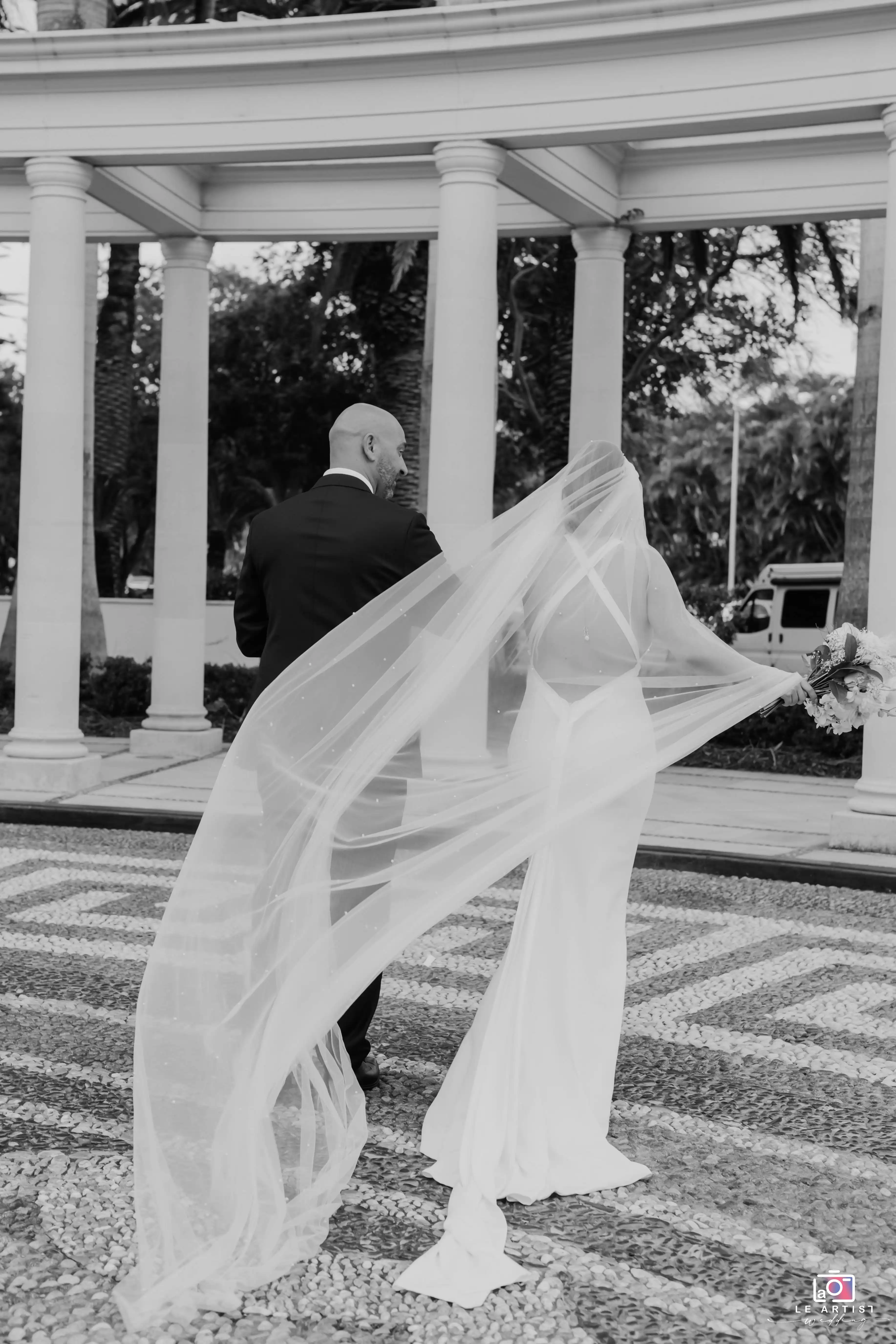 The newlywed couple walks gracefully under a grand colonnade, the bride’s veil flowing elegantly in the breeze. Le Artist Wedding