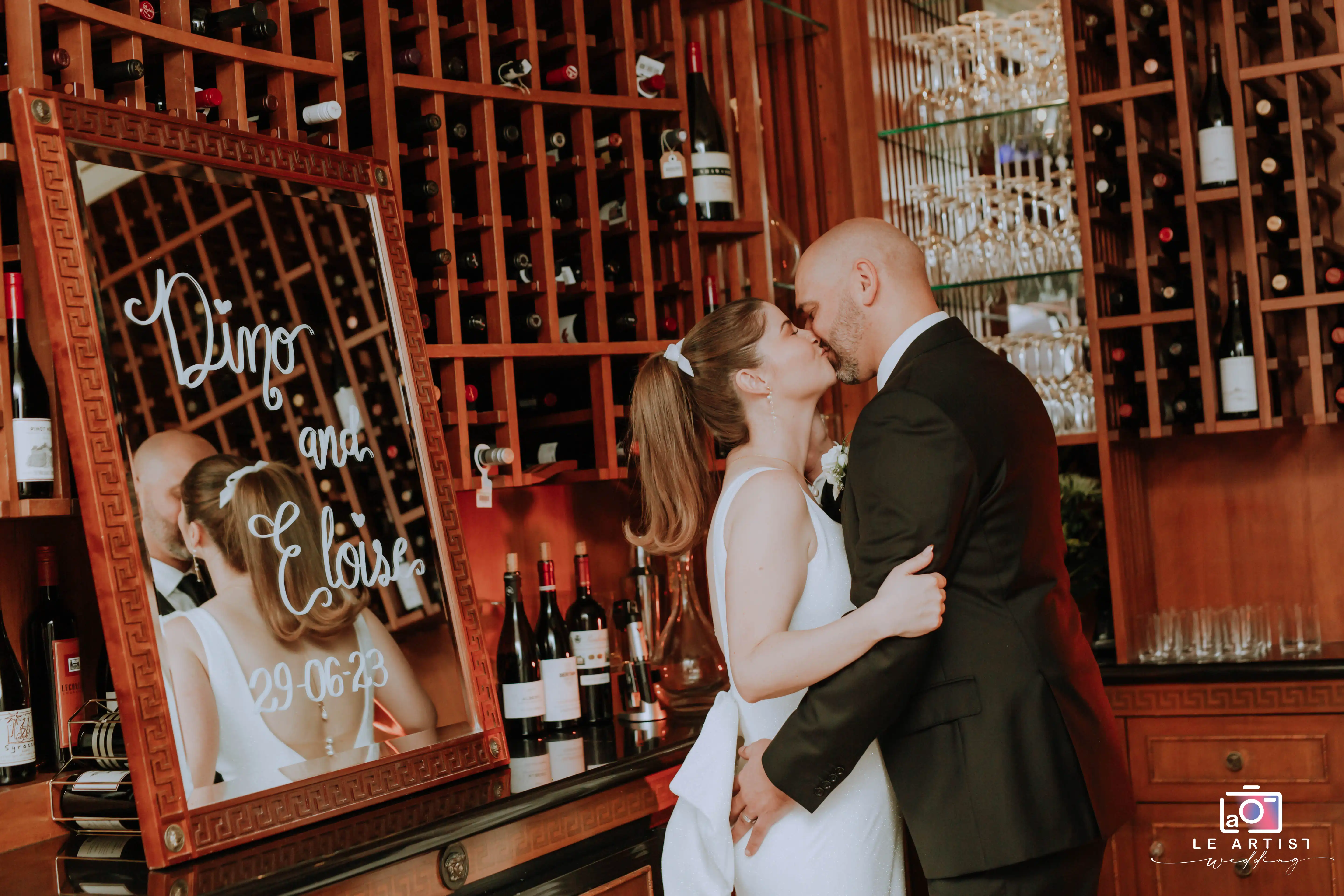 The bride and groom share a romantic kiss in an elegant wine cellar during their bridal photoshoot, captured beautifully beside a personalized mirror. Le Artist Wedding