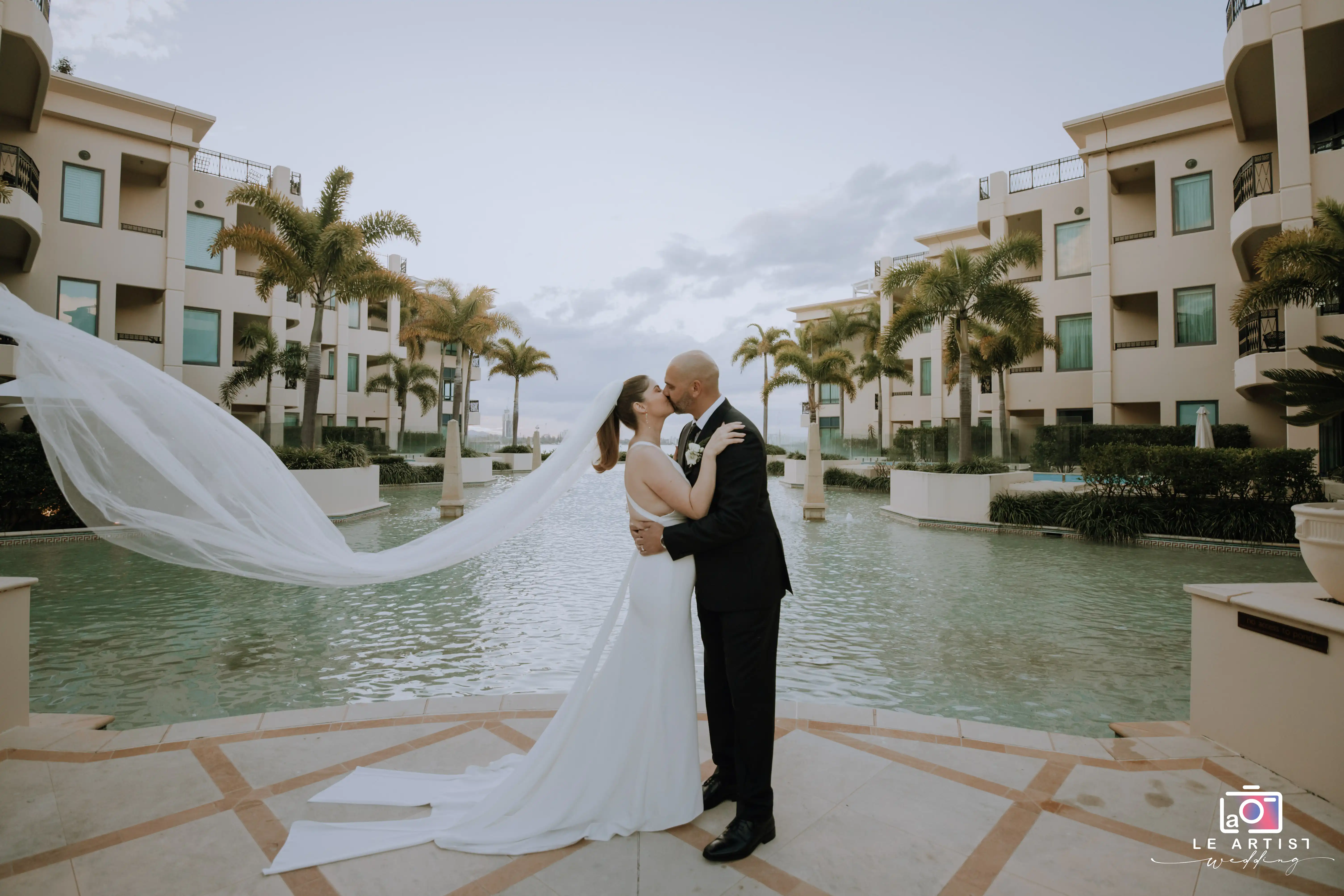 The bride and groom share a romantic kiss by a serene water feature, surrounded by elegant architecture and palm trees, capturing a perfect moment of their wedding day. Le Artist Wedding