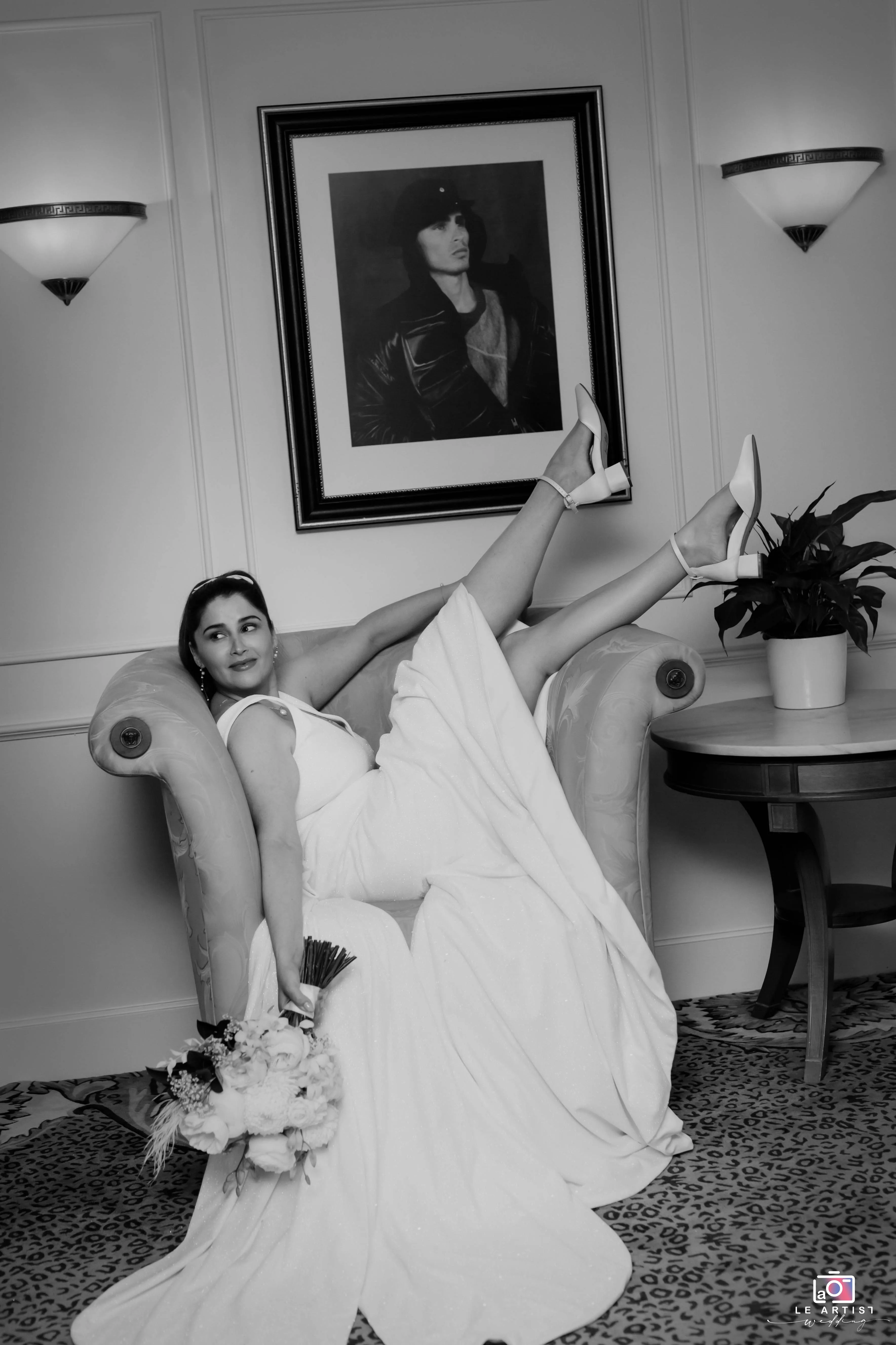 The bride strikes a playful pose on an elegant armchair, showcasing her gown and bouquet in an intimate and artistic setting. Le Artist Wedding