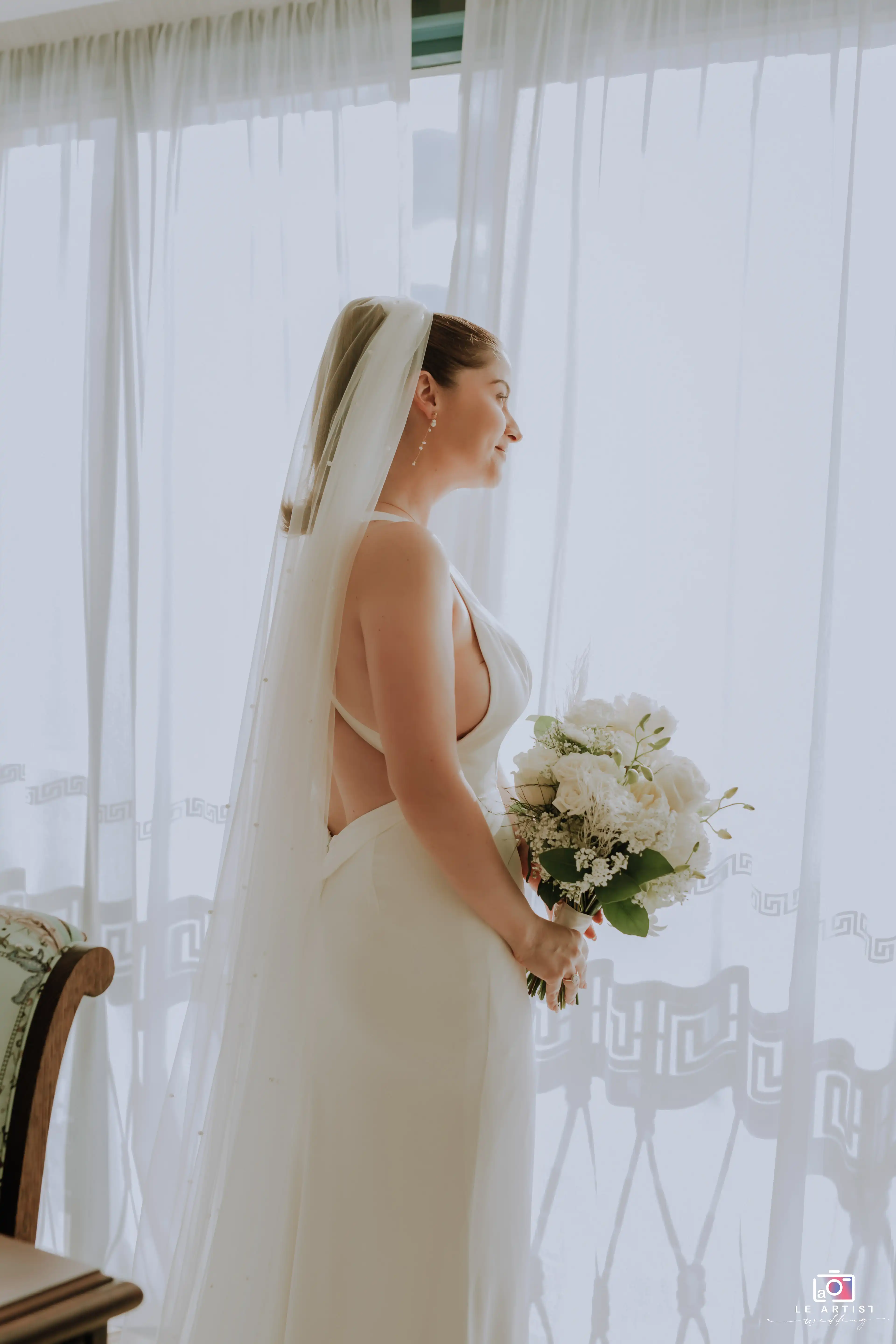 The bride stands gracefully by the window, holding her bouquet, as natural light illuminates her elegant gown and veil. Le Artist Wedding