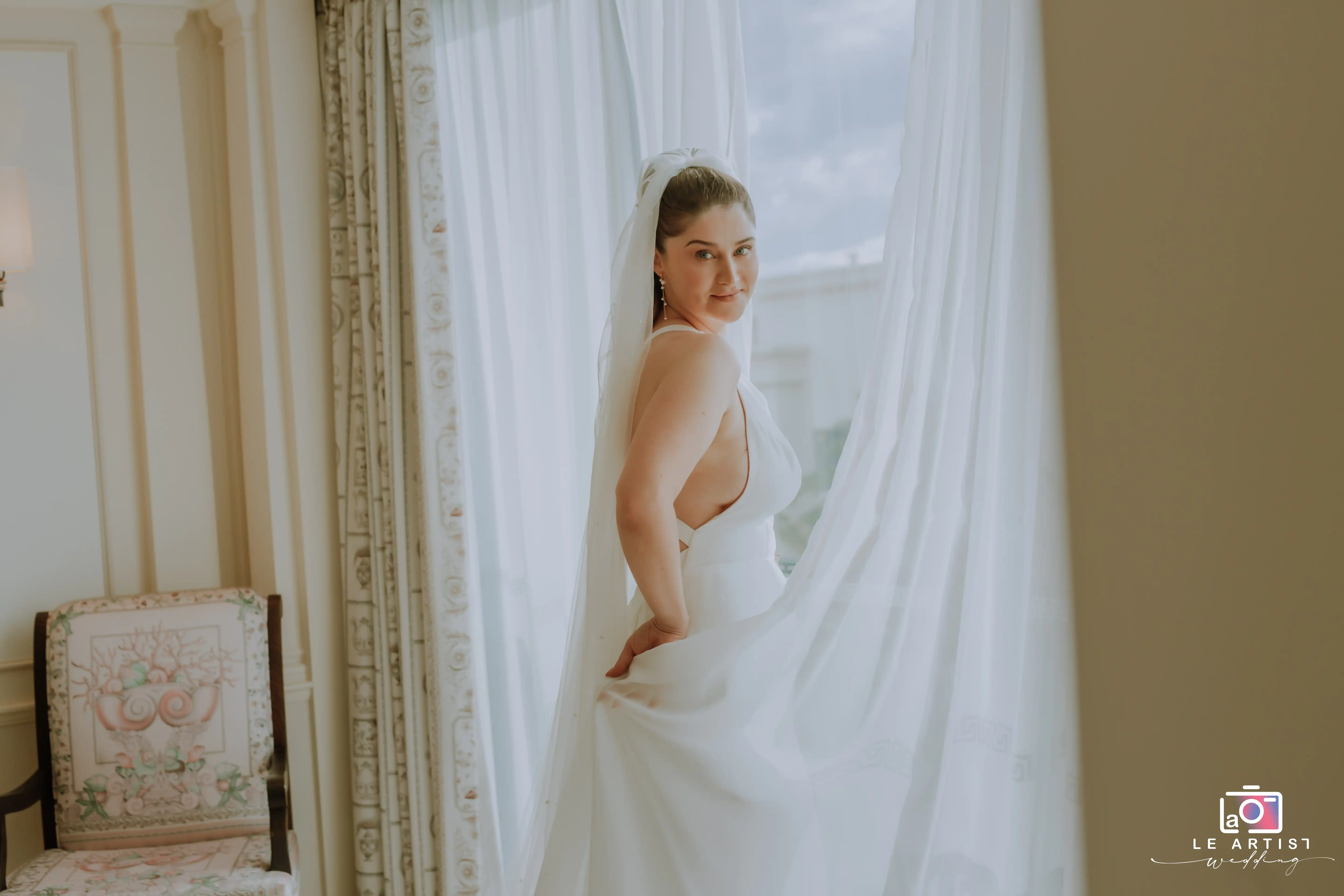 The bride poses gracefully by a window, her elegant gown catching the light in an intimate indoor photoshoot. Le Artist Wedding