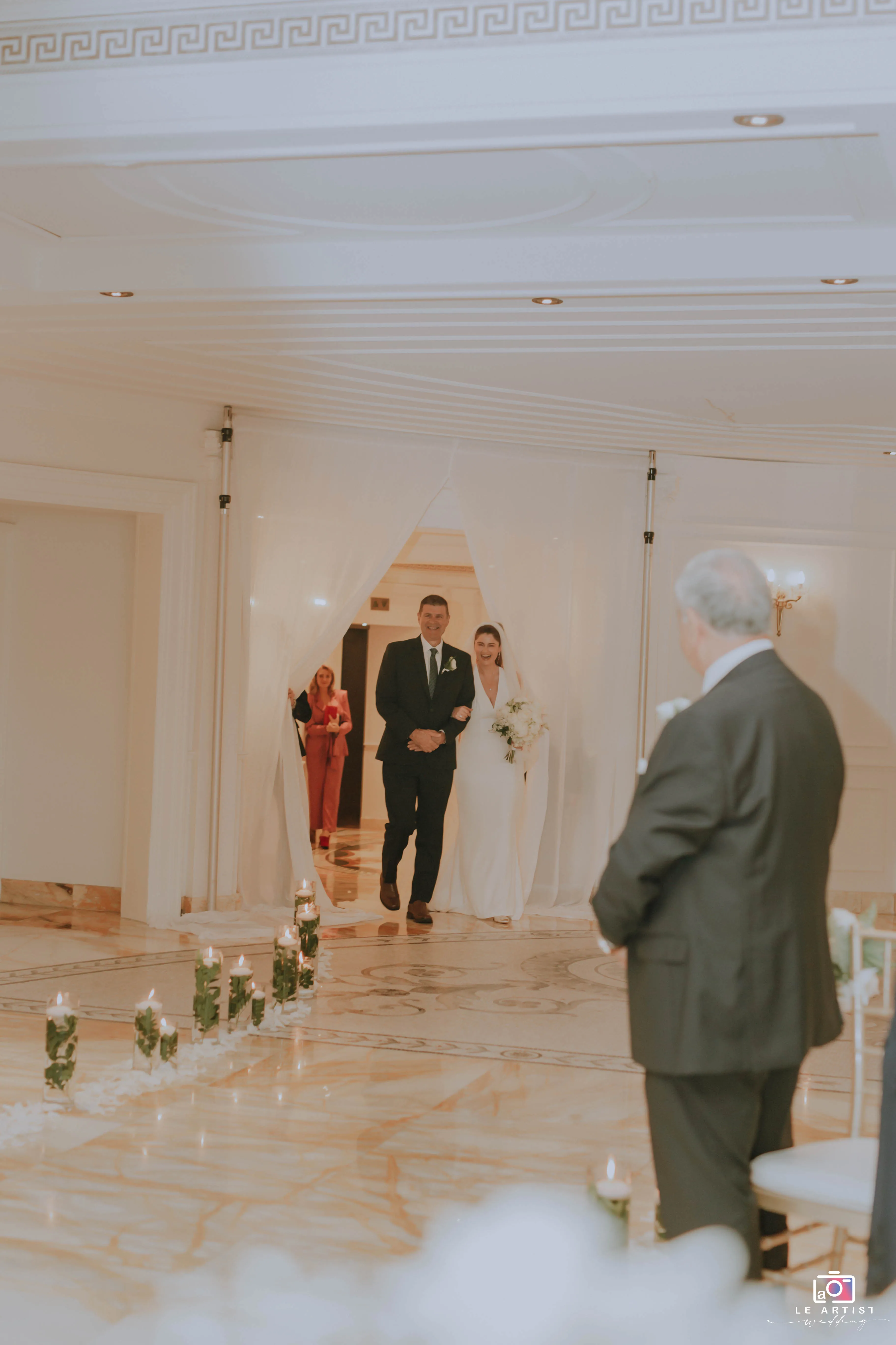 The bride makes her entrance during the wedding ceremony, walking down the aisle accompanied by a loved one. Le Artist Wedding