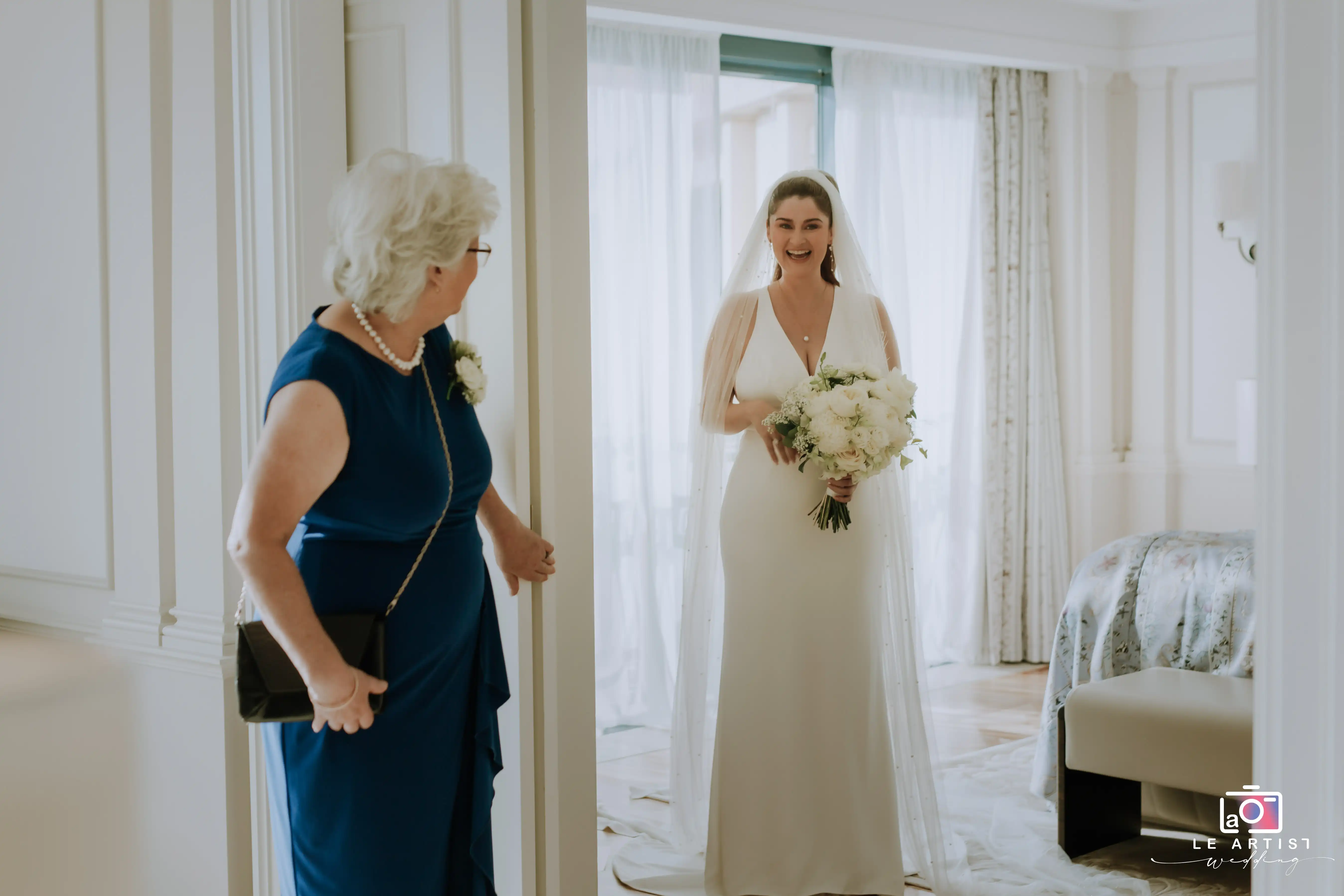 The bride shares a joyful moment with a loved one, showcasing her elegant wedding dress and bouquet as she gets ready for the big day. Le Artist Wedding