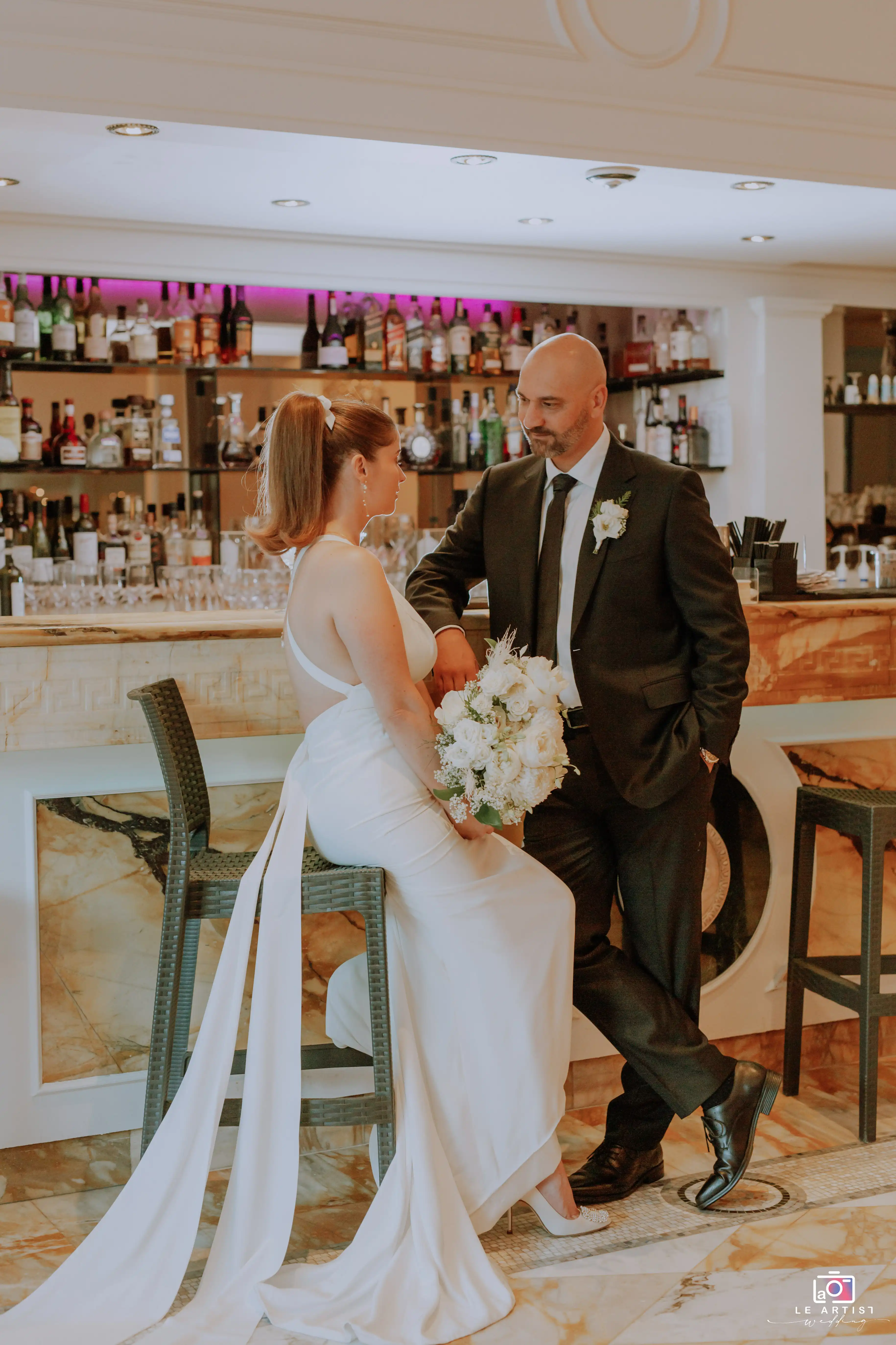 The bride and groom share a moment at the bar in an elegant indoor photoshoot. Le Artist Wedding