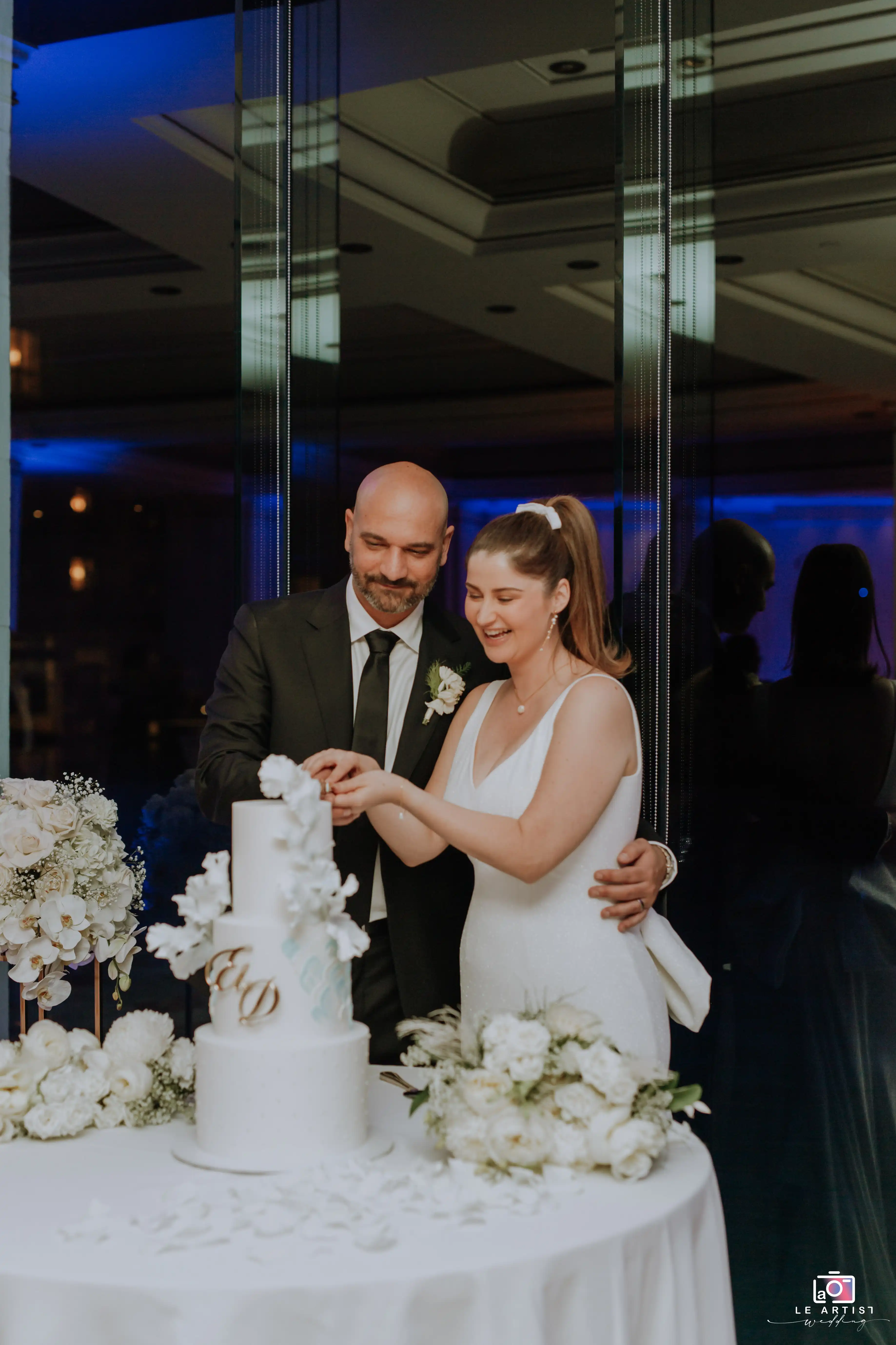 The bride and groom joyfully cut their wedding cake, surrounded by elegant floral arrangements, during the reception celebration. Le Artist Wedding