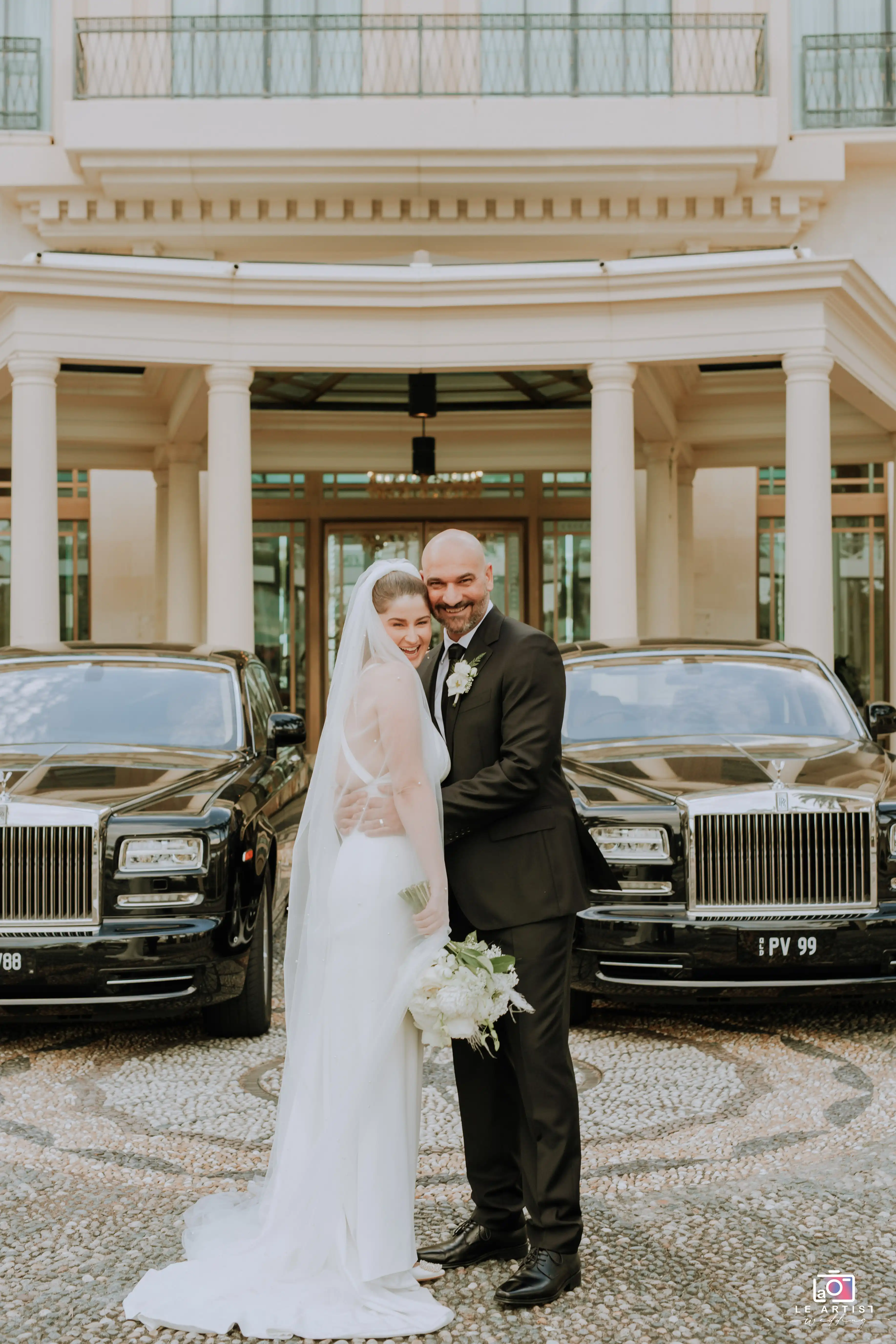 The bride and groom elegantly pose in front of classic cars, capturing a moment of joy and elegance. Le Artist Wedding