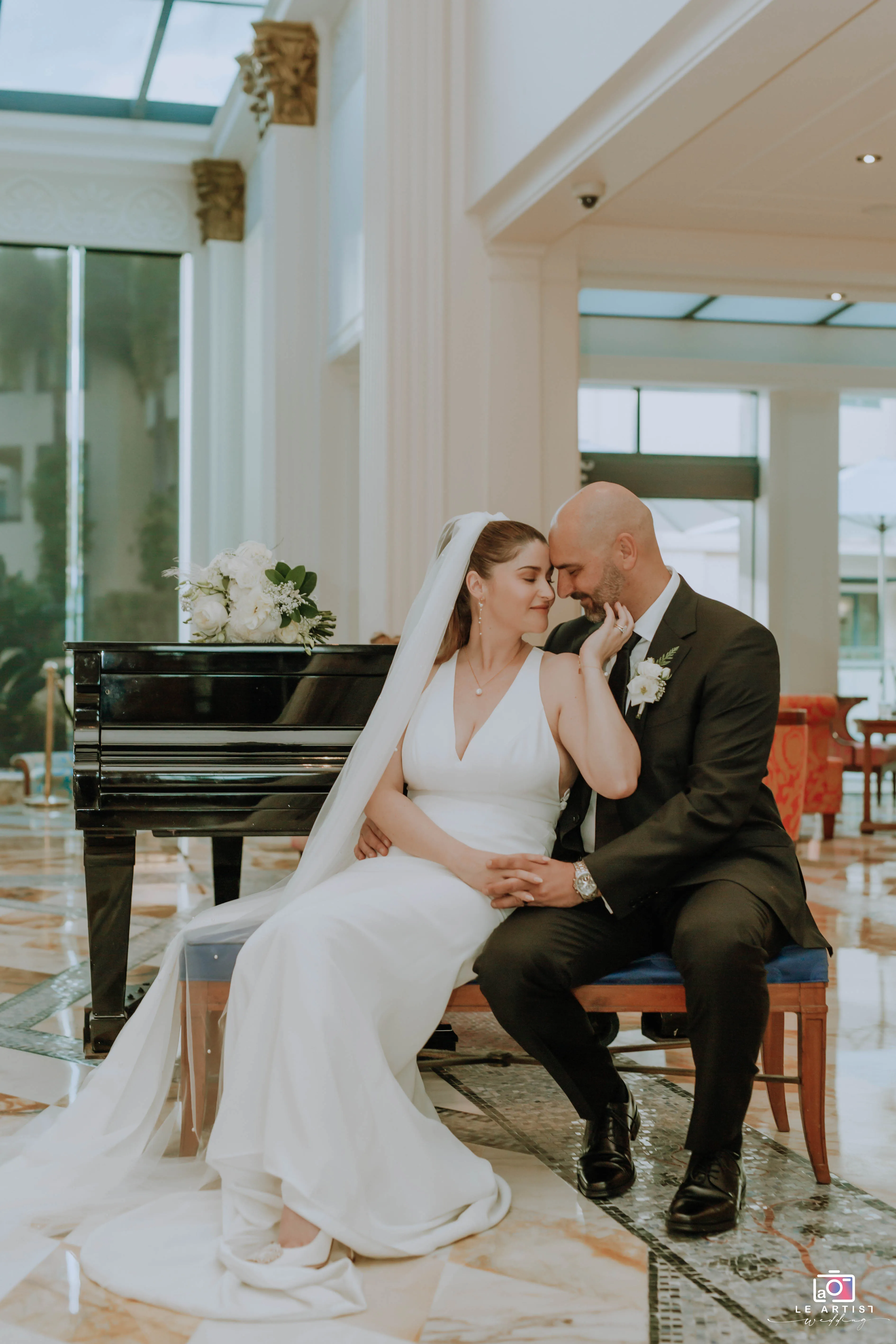 The bride and groom share an intimate moment seated by a grand piano, capturing their love and elegance in an indoor photoshoot. Le Artist Wedding