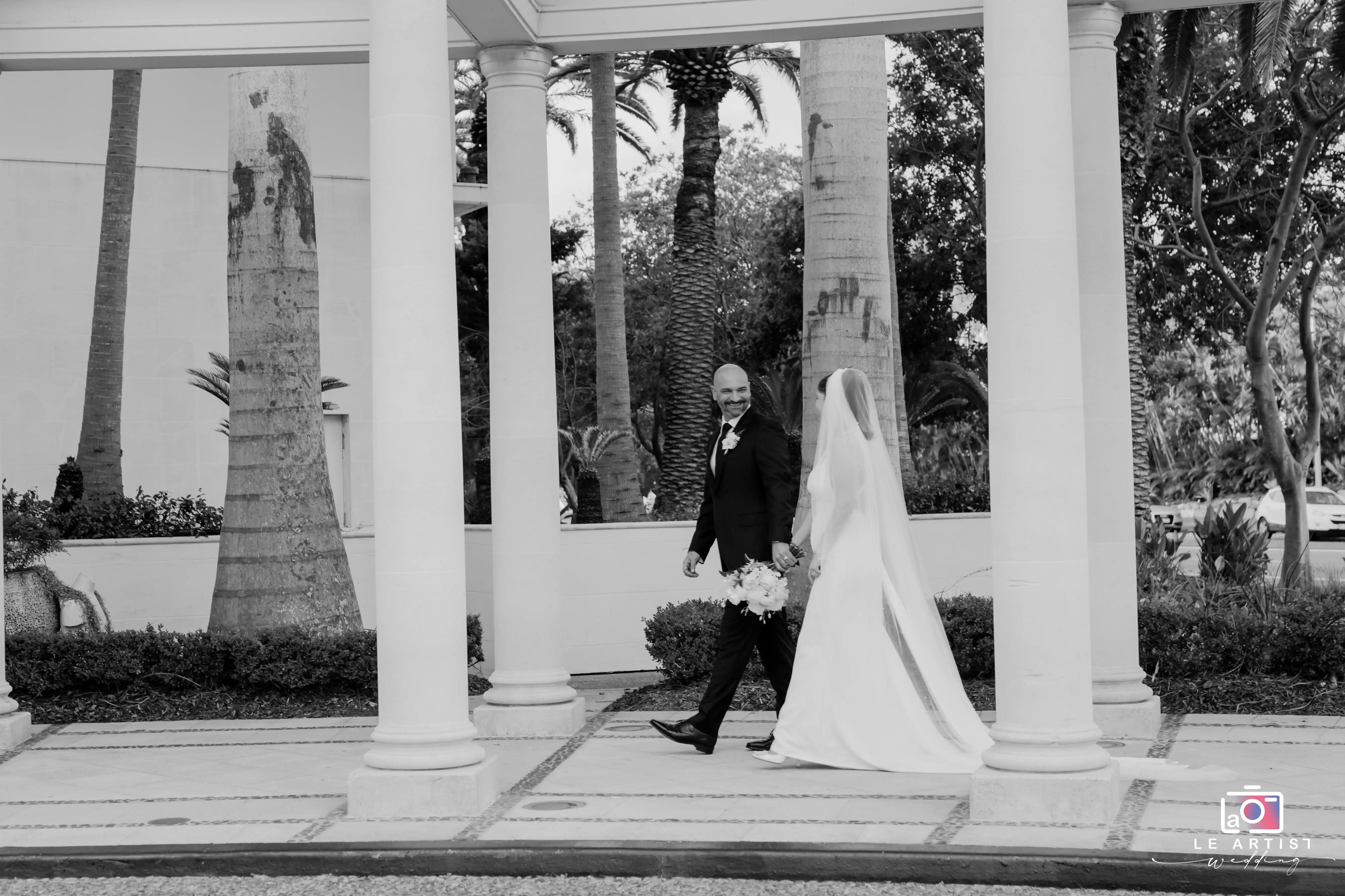 The bride and groom share a joyful moment during their outdoor photoshoot, holding hands and walking amidst elegant columns. Le Artist Wedding