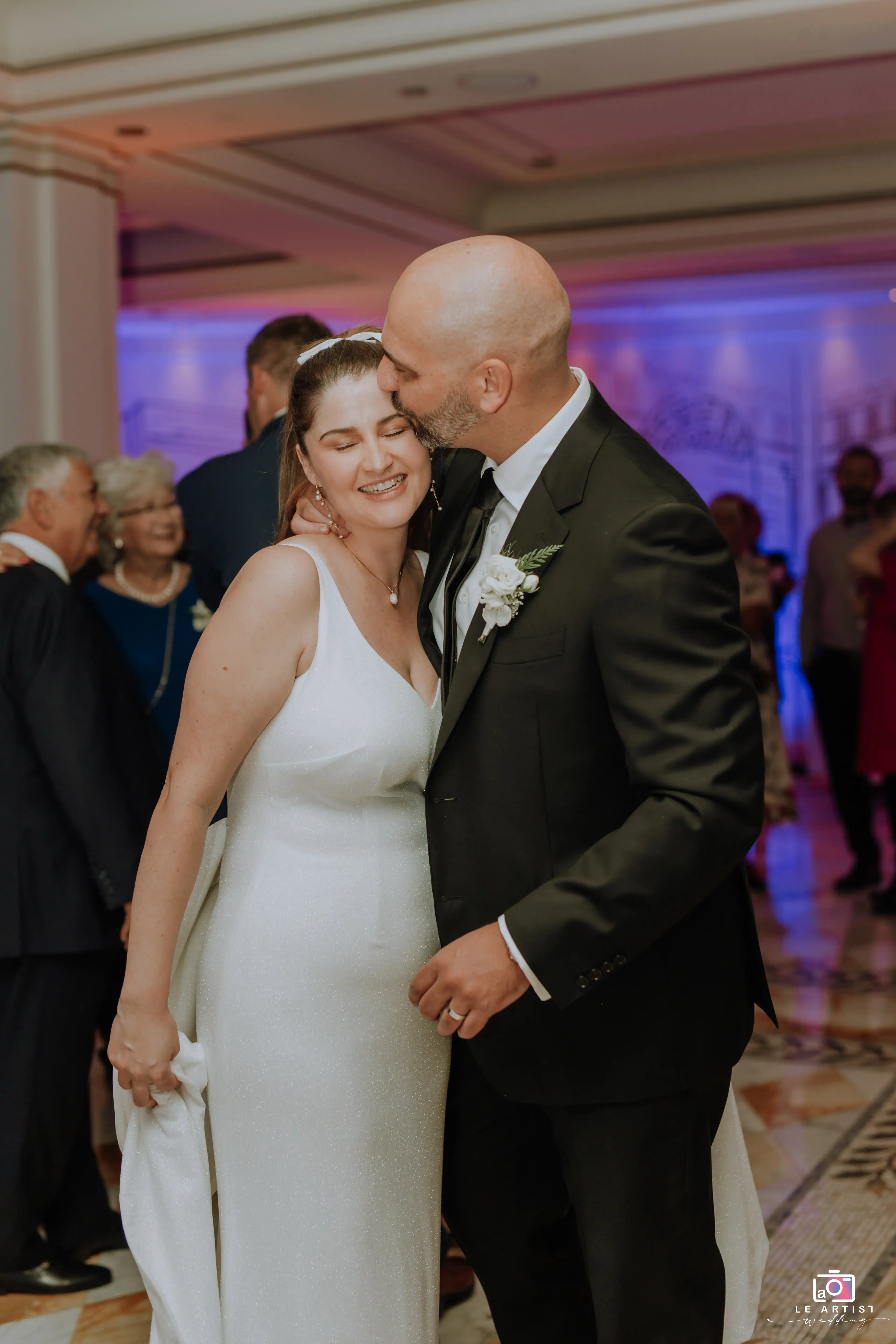 The bride and groom share a joyful moment during the wedding reception, surrounded by their guests. Le Artist Wedding