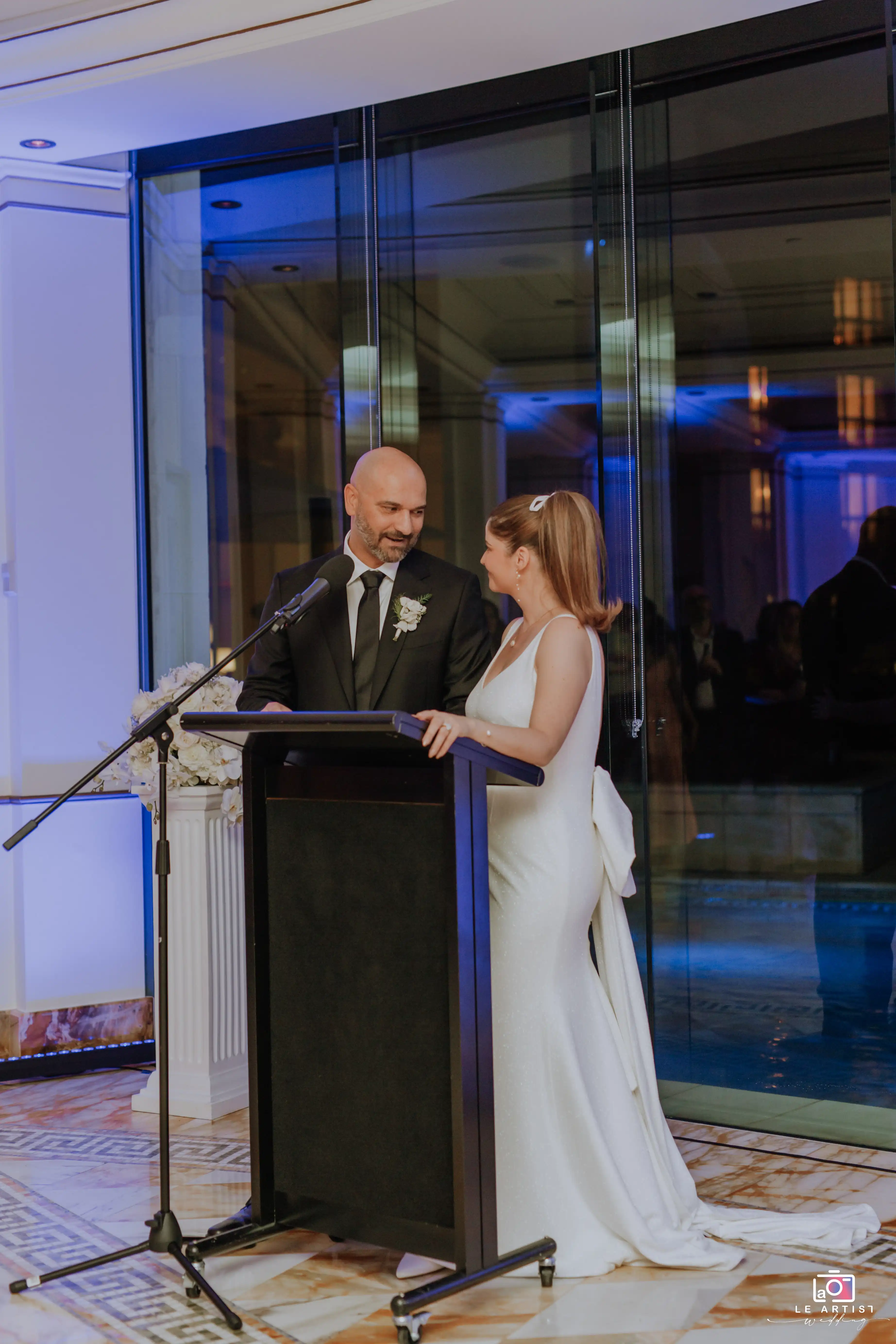 The bride and groom share a heartfelt moment at the microphone during the reception, surrounded by the warm ambiance of their celebration. Le Artist Wedding