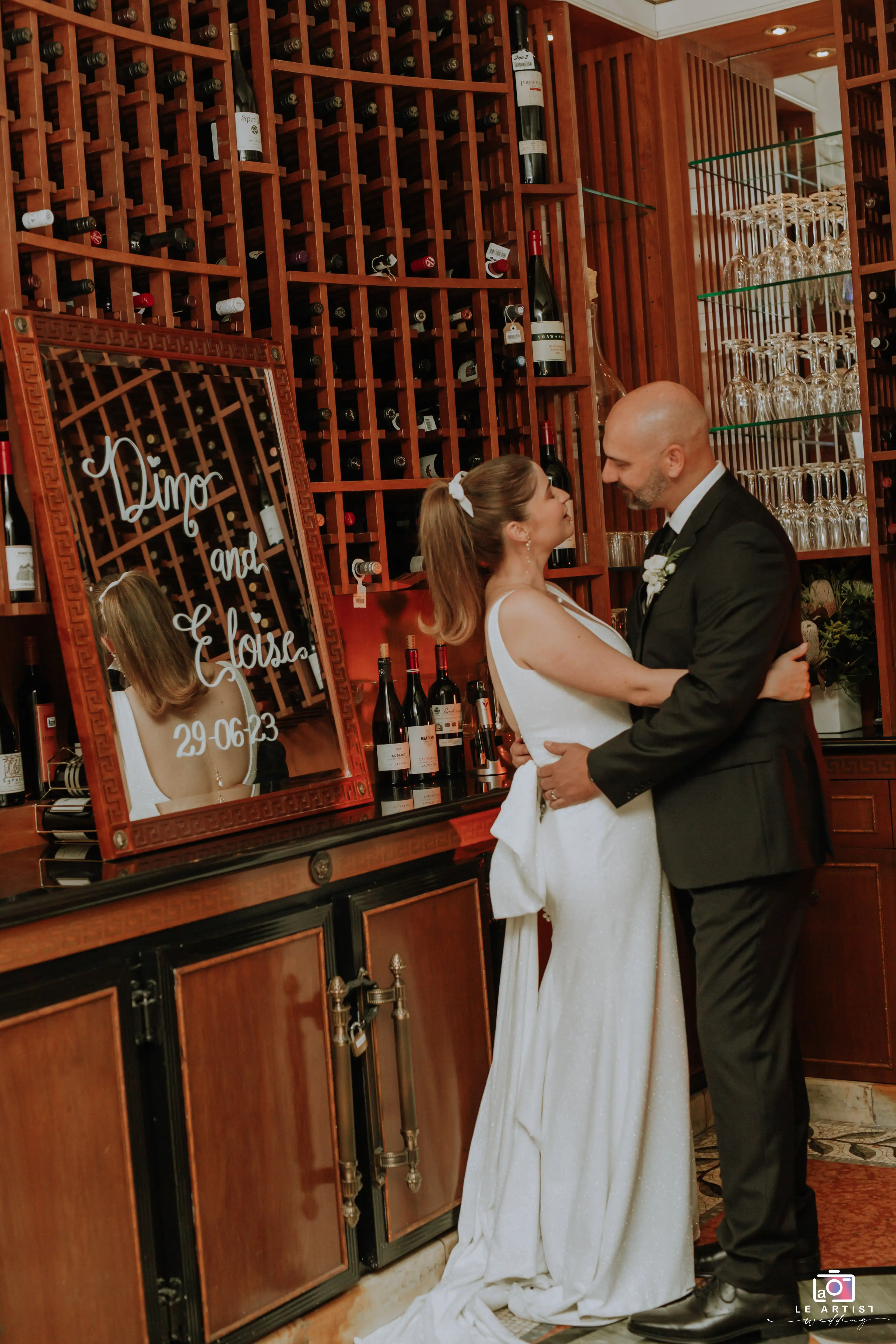 The bride and groom share an intimate moment in a stylish setting, surrounded by wine bottles, creating a warm and romantic atmosphere. Le Artist Wedding
